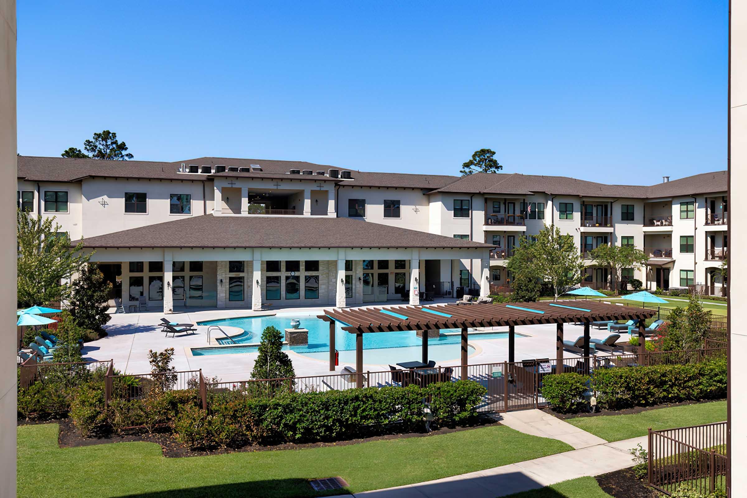 A view of a modern apartment complex featuring a swimming pool surrounded by lounge chairs and umbrellas, with well-maintained landscaping and a clear blue sky in the background.