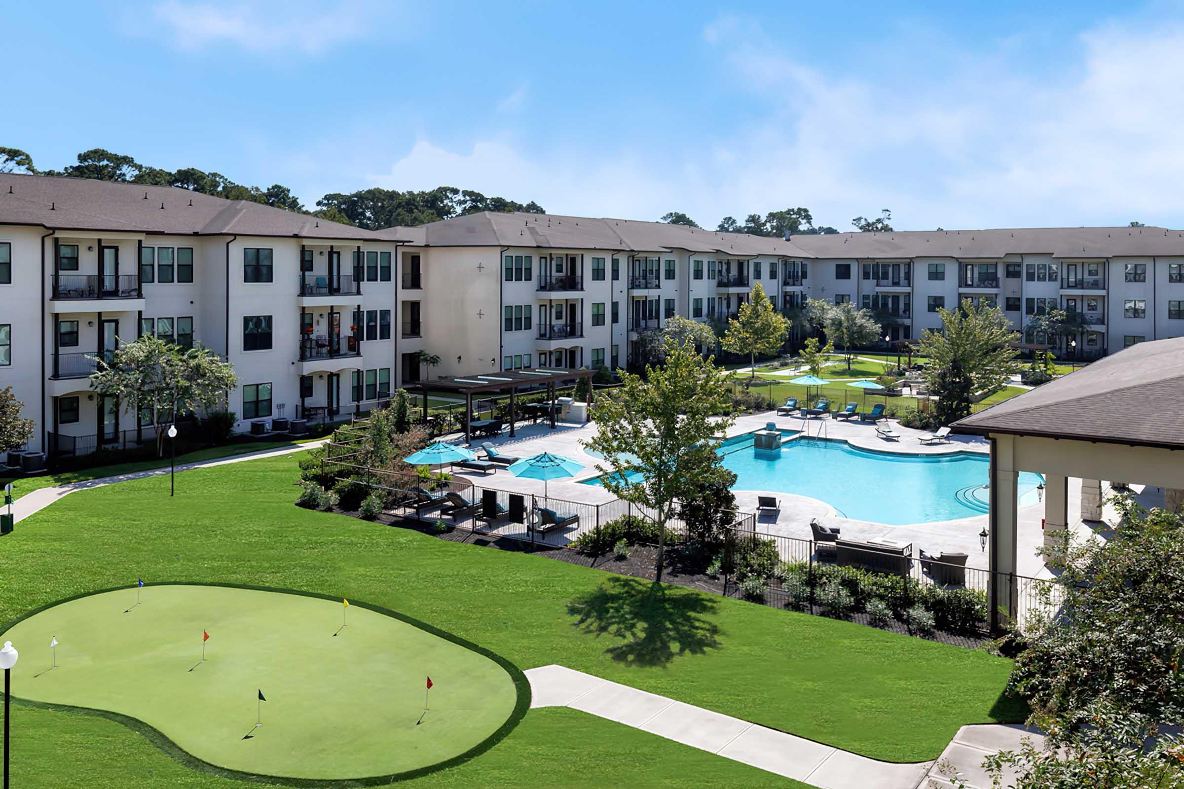 View of an apartment complex featuring a landscaped courtyard with a swimming pool, lounge chairs, and a putting green in the foreground. Several buildings surround the pool area, and trees provide greenery. Blue sky is visible above.