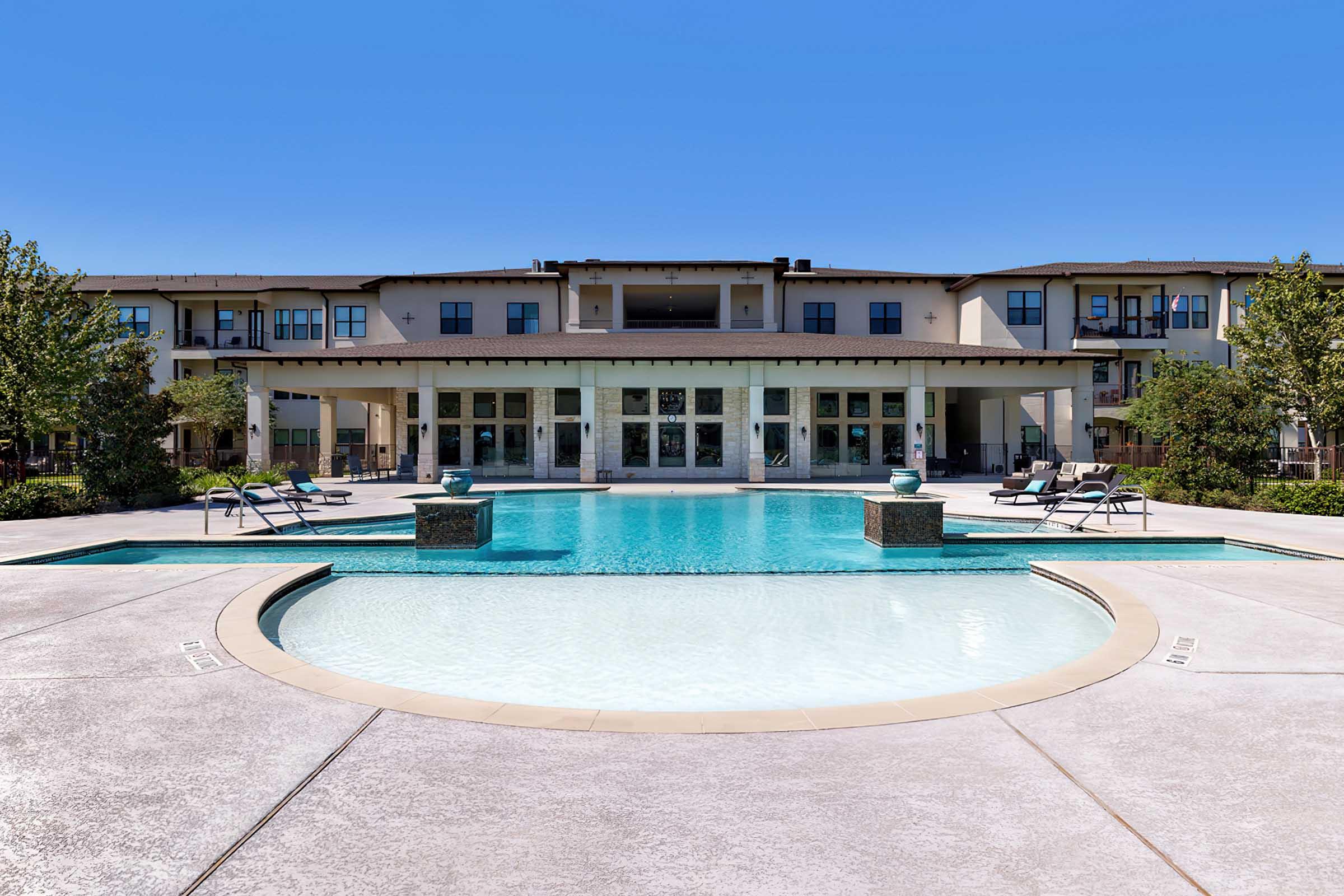 A modern outdoor swimming pool with a circular design, surrounded by lounge chairs and palm trees. The pool area is in front of a stylish multi-story building with large windows and a patio, set against a clear blue sky.