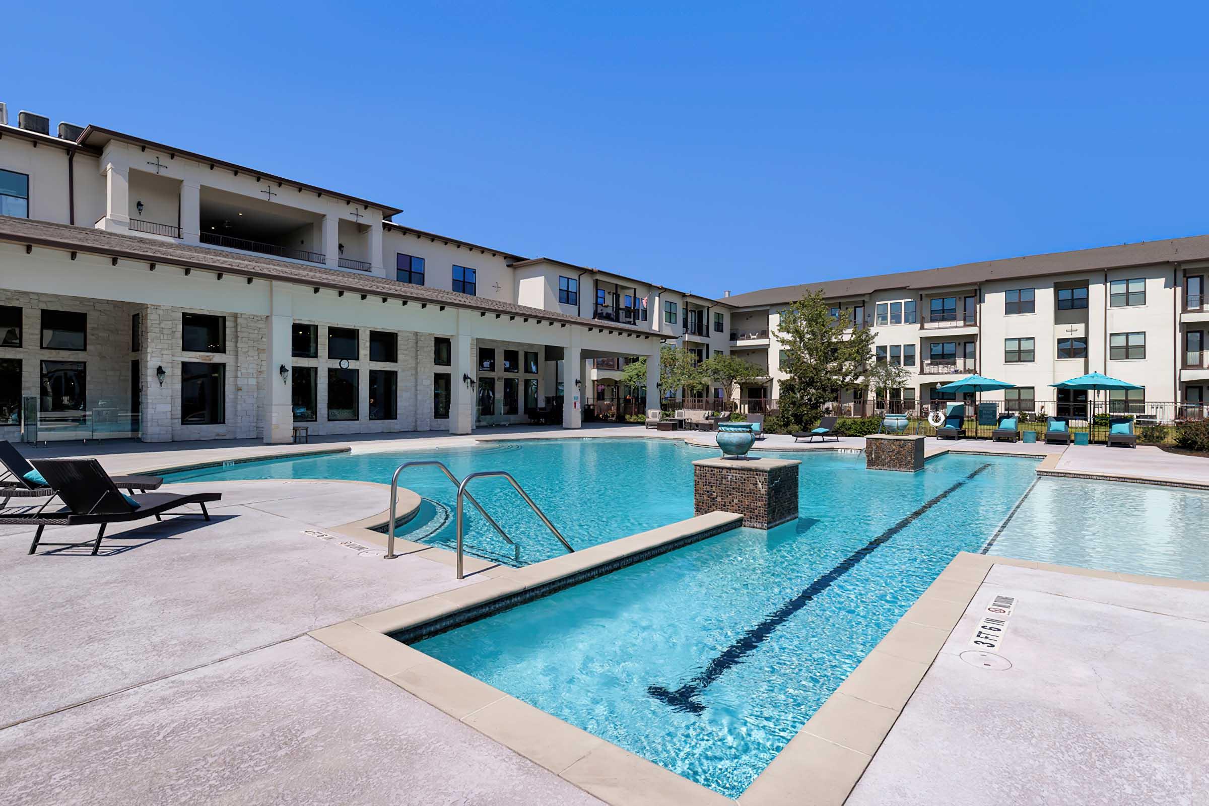 A modern apartment complex featuring a large outdoor swimming pool surrounded by lounge chairs, with a sunny blue sky and green landscaping in the background.