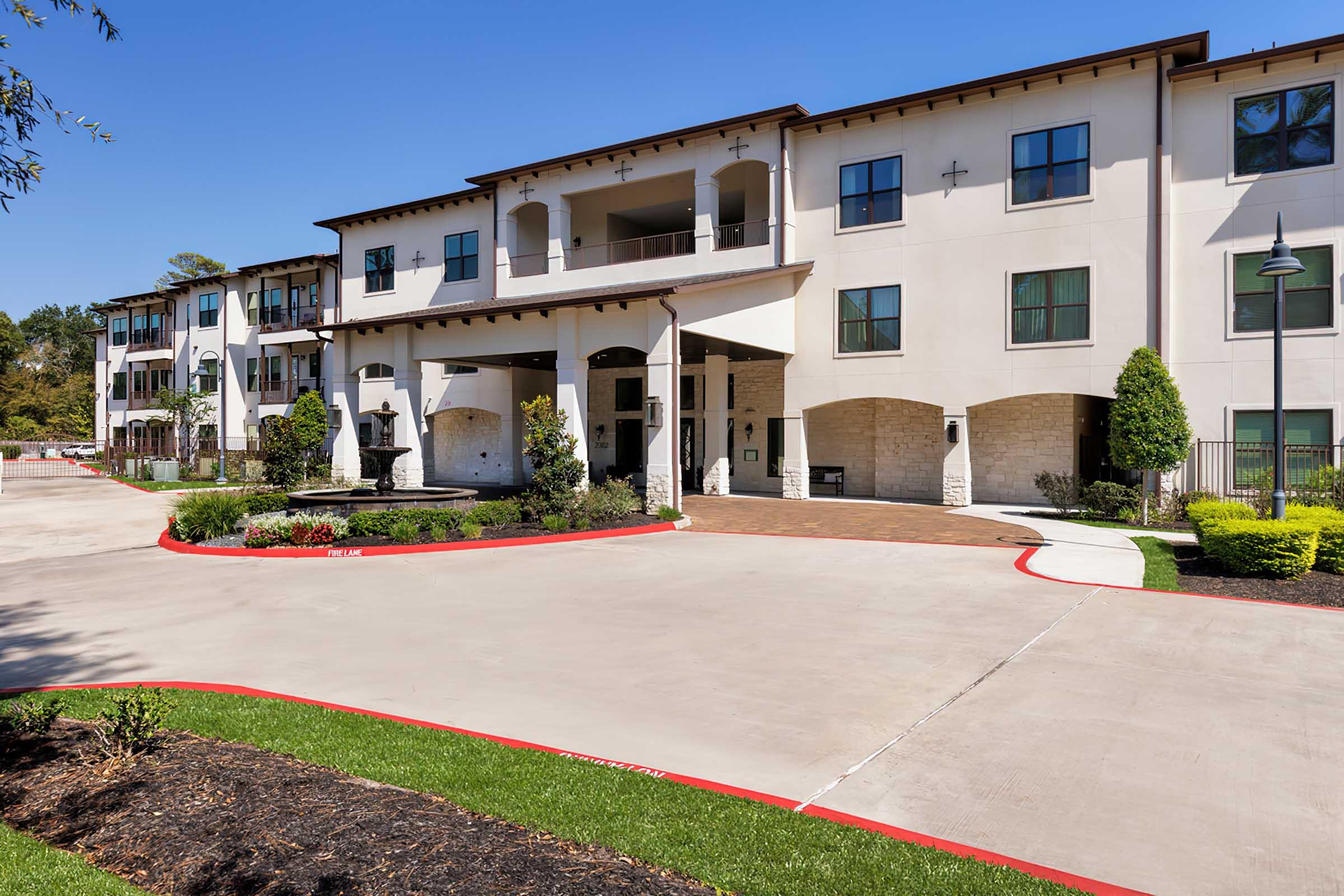 A modern, multi-story residential building with a welcoming entrance, featuring stone columns, a landscaped driveway, and decorative shrubs. Bright blue sky overhead.