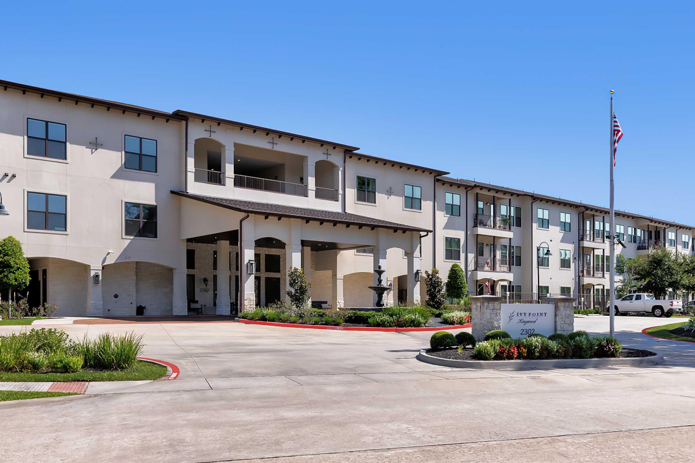 A modern residential building with multiple stories, featuring a welcoming entrance and landscaped surroundings. The structure has a light-colored facade and a covered entryway. An American flag is displayed prominently near the entrance, and there are parked cars visible in the vicinity.