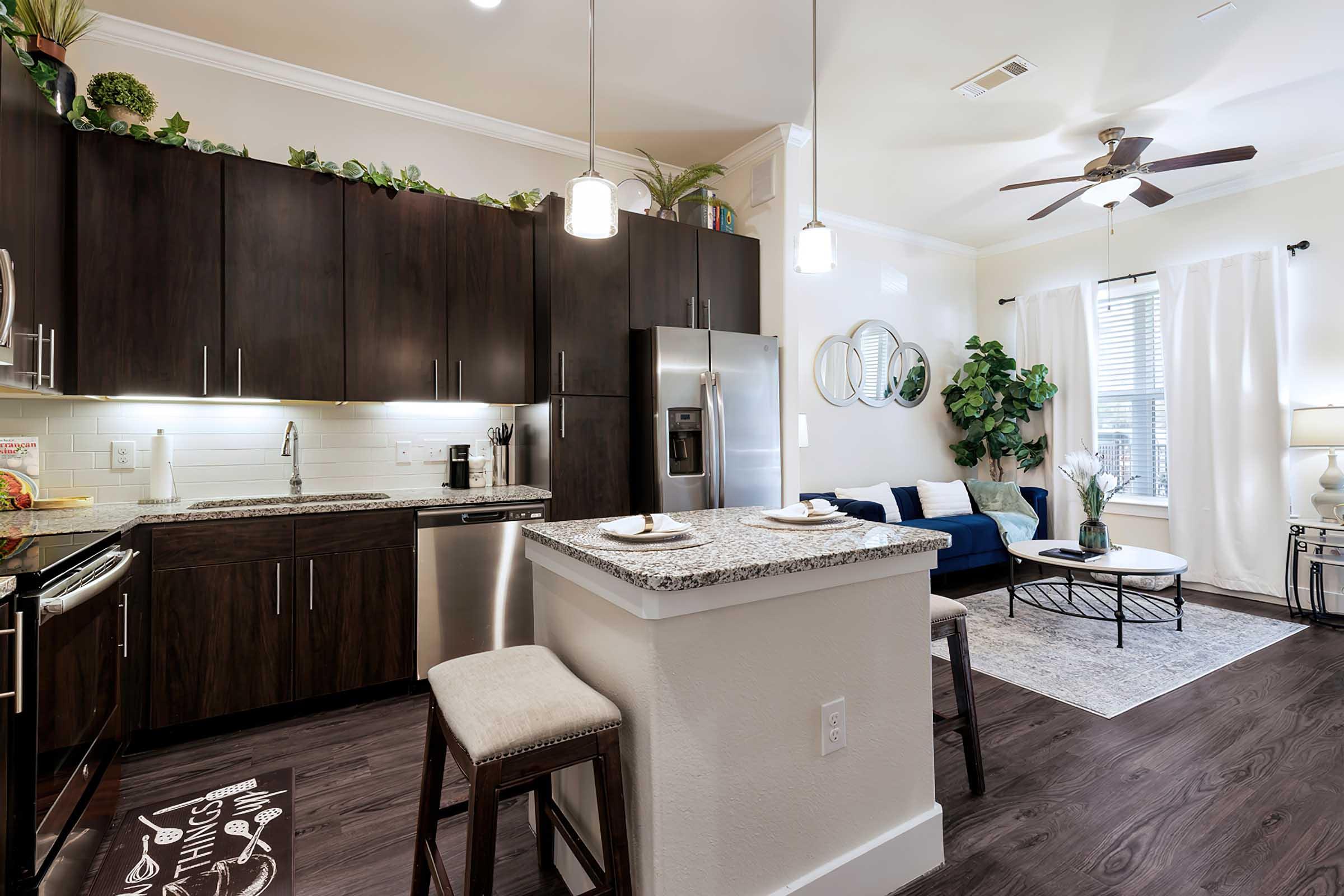 Modern kitchen and living area featuring dark wood cabinets, stainless steel appliances, a granite countertop island with bar stools, and a cozy seating area with a sofa and decorative plants. Bright natural light comes through the windows, complemented by light-colored curtains.