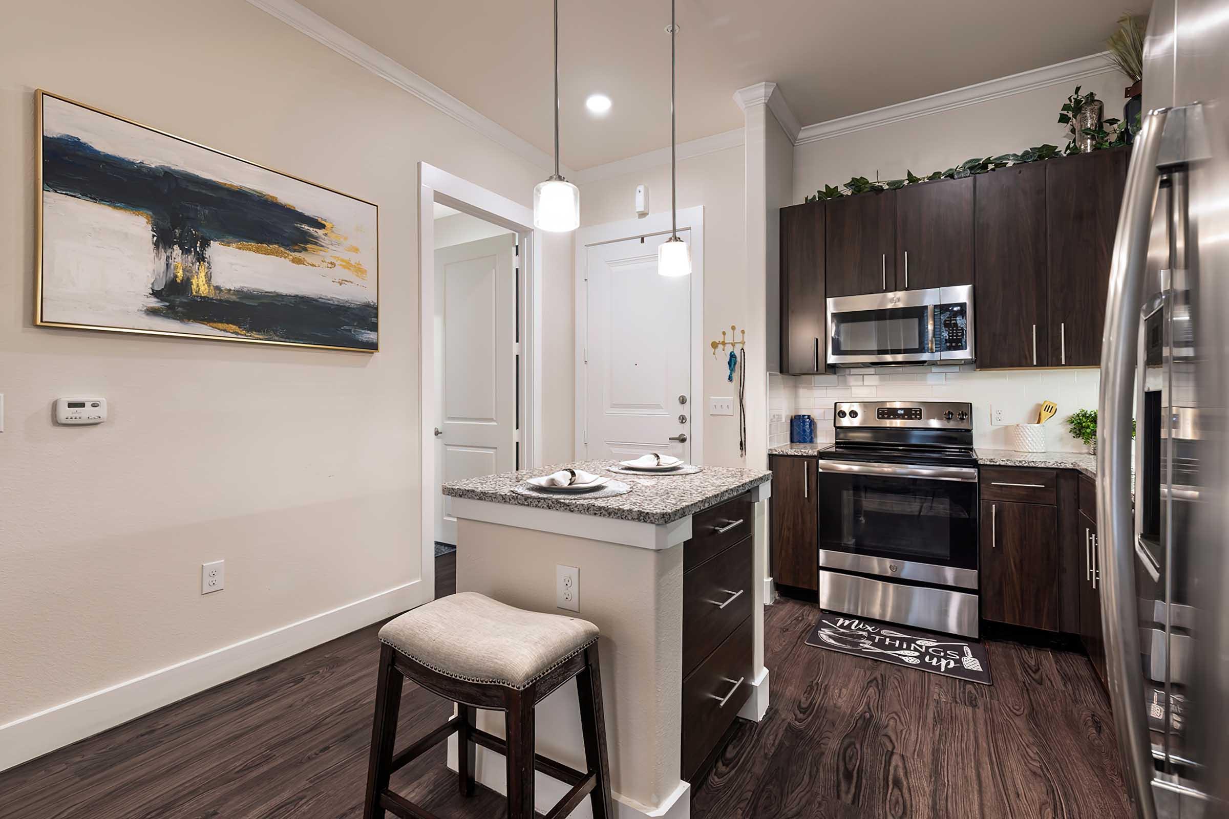 Modern kitchen featuring dark wood cabinets, stainless steel appliances, and a granite island with seating. Pendant lights hang above the island, and there is a piece of abstract artwork on the wall. The flooring is hardwood, and the overall color scheme is neutral with touches of blue and black.