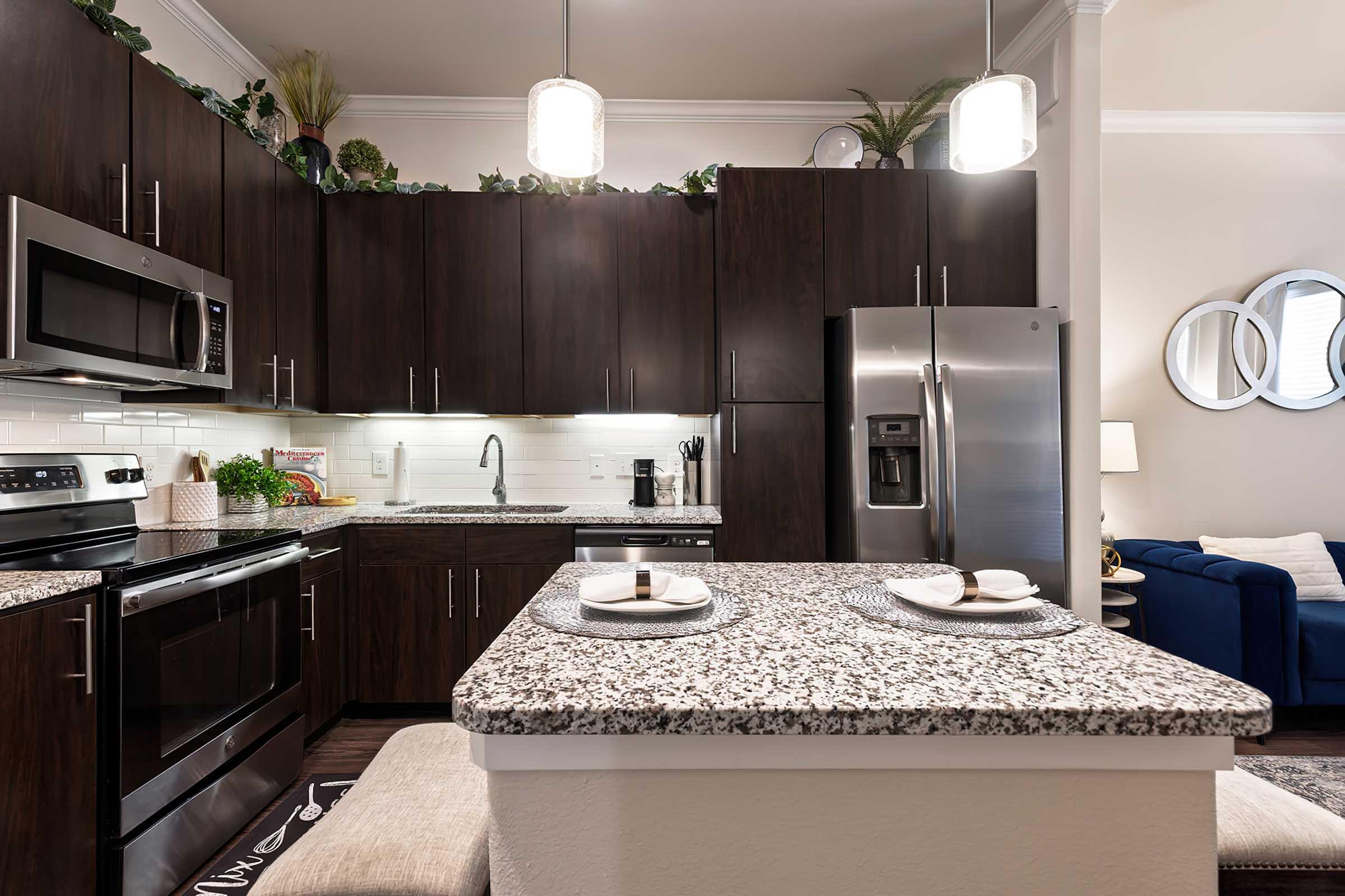 Modern kitchen with dark wood cabinets, granite countertops, stainless steel appliances, and decorative plants. Features an island with two place settings, and a cozy living area visible in the background.