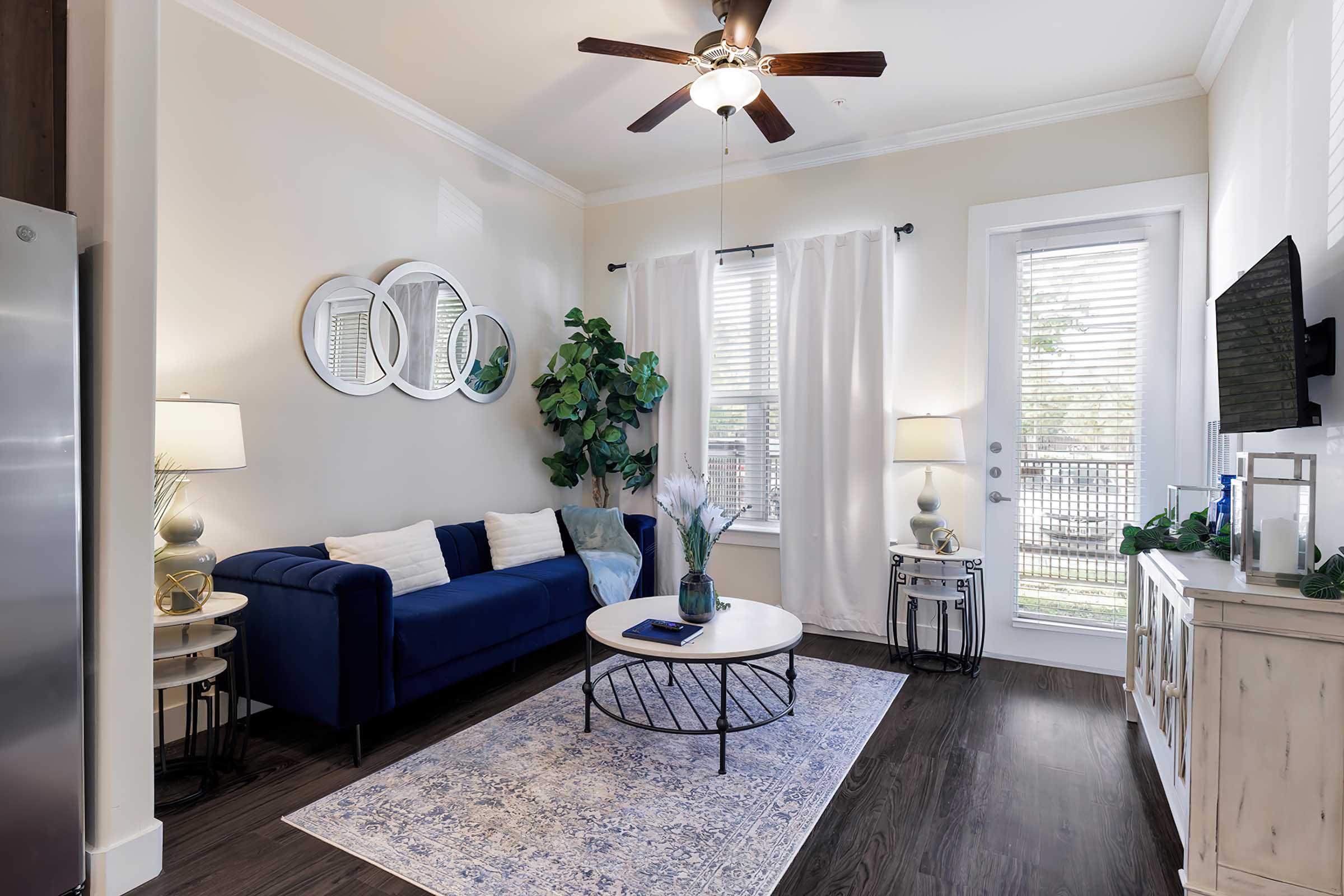 A stylish living room featuring a blue sofa, a round coffee table on a light-colored rug, and a large window with sheer curtains. There are two lamps on either side of the sofa, a potted plant, and a television on the wall opposite the sofa. The decor is modern and inviting, with a ceiling fan adding to the ambiance.