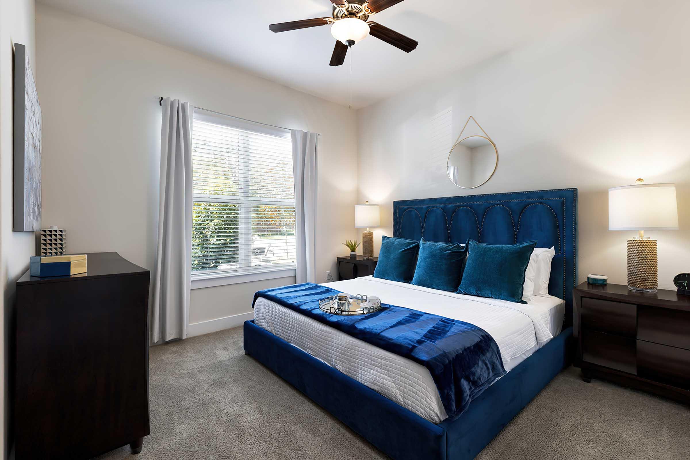 A cozy bedroom featuring a blue upholstered bed with three decorative pillows, a plush blue throw blanket, and light-colored walls. The room has a ceiling fan, two bedside lamps on dark wood nightstands, and a window with sheer curtains allowing natural light to fill the space. A decorative wall mirror and a piece of artwork can be seen, along with a dresser beside the bed.