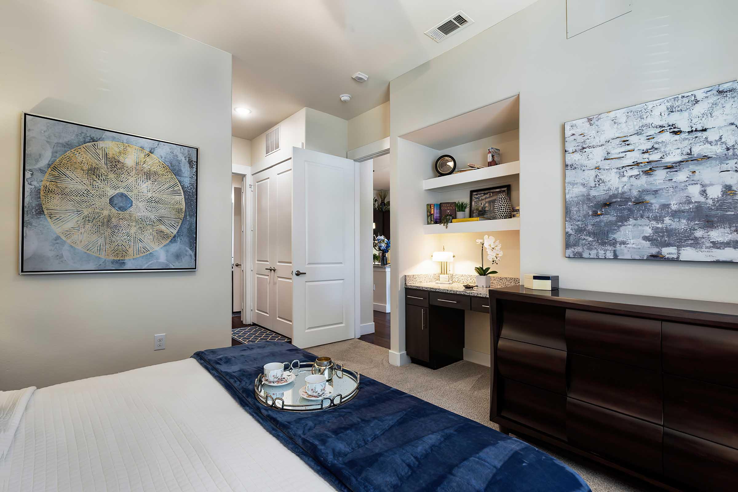 A cozy bedroom featuring a neatly made bed with a blue throw blanket, decorative trays with drinks, and modern artwork on the walls. A door leads to another room, and there is a small desk area with a lamp and decorative items. Soft lighting creates a welcoming atmosphere.