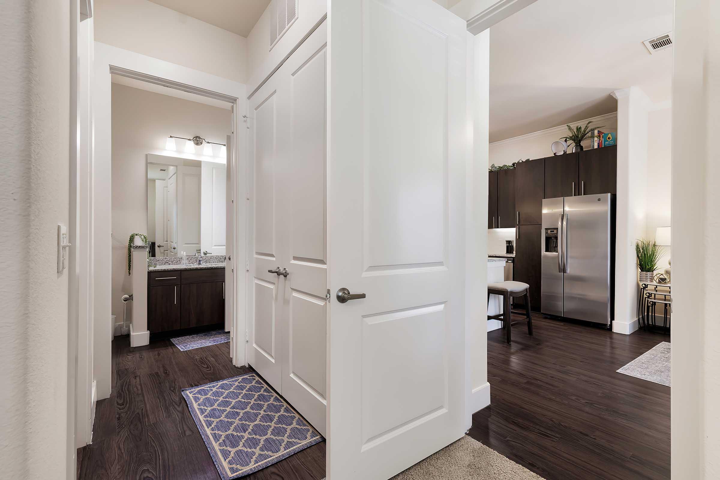 Interior view of a modern apartment hallway, featuring an open doorway leading to a bathroom with a sink and mirror, and another doorway opening into a kitchen area with dark cabinetry and stainless steel appliances. The floor is dark wood, and there are area rugs on the floor.