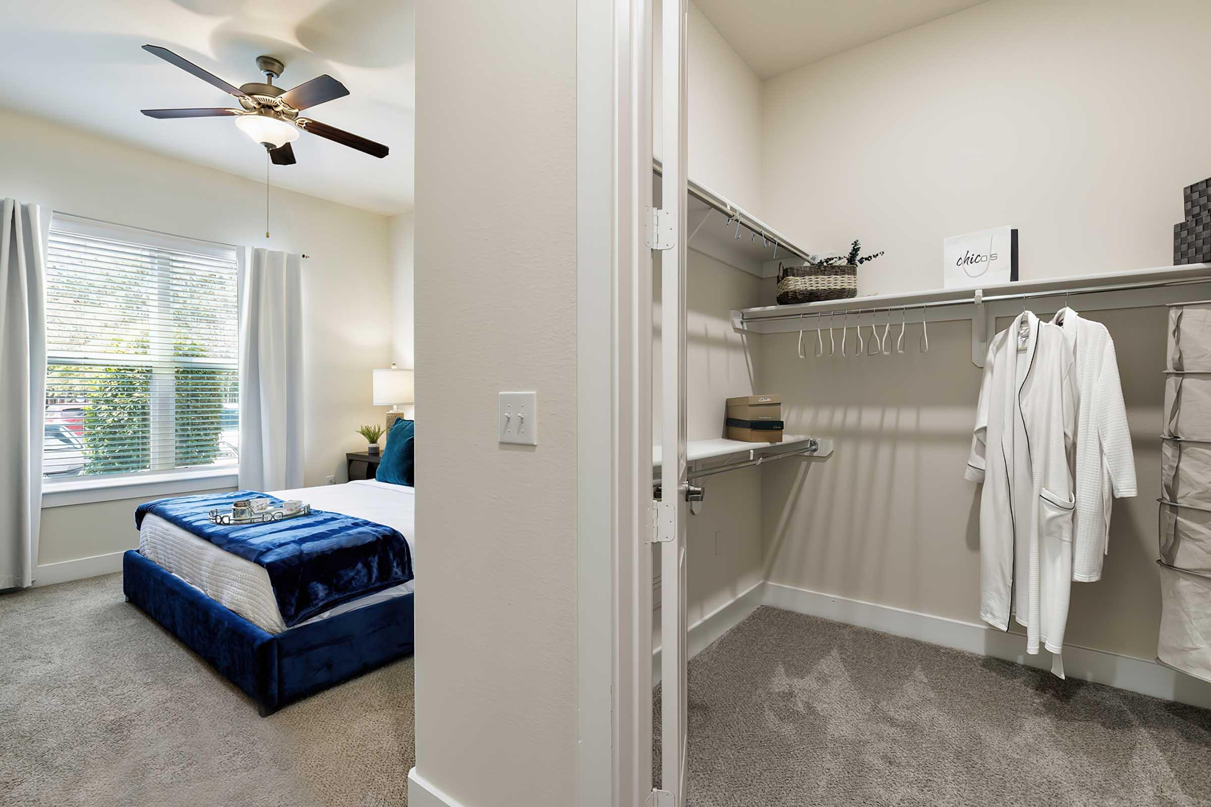 A well-lit bedroom with a bed covered in blue bedding, a nightstand with a lamp, and a window with sheer curtains. Adjacent to the bedroom is a closet featuring a rack with hangers, a hanging robe, and storage baskets. The carpeted floor adds warmth to the space.