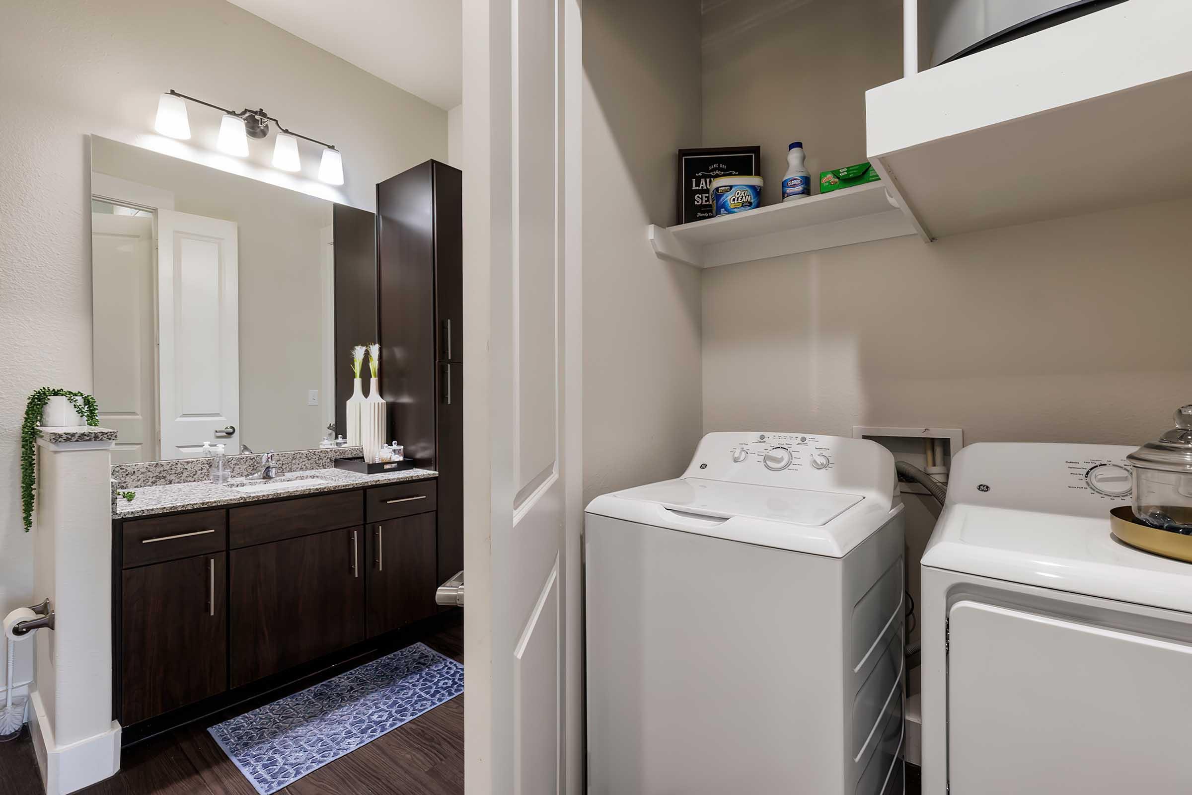 A modern bathroom featuring a double vanity with dark cabinets, a mirror, and decorative items. To the side, there is a laundry area with a washer and dryer. The flooring is dark, and there is a small rug in front of the vanity.