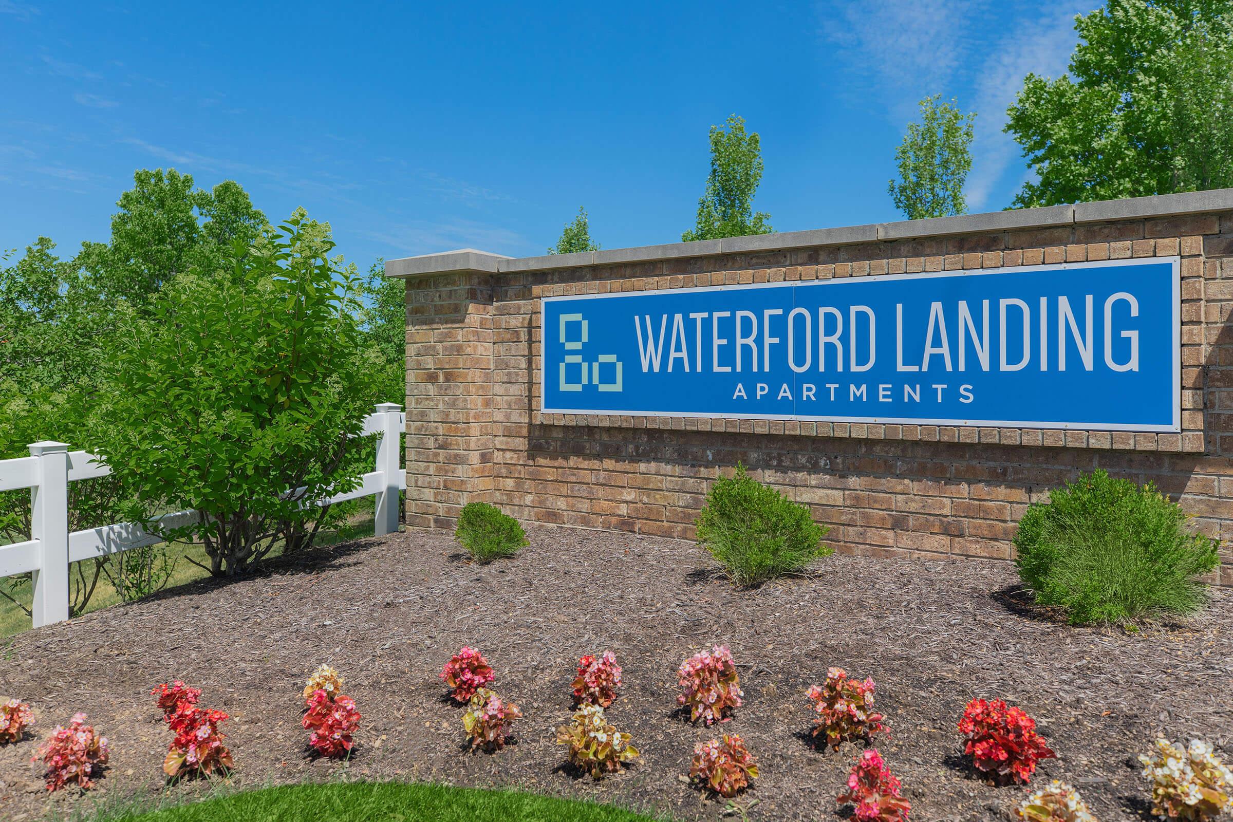 Sign for Waterford Landing Apartments featuring the name prominently displayed on a blue background, surrounded by manicured landscaping with flowering plants and trees under a clear blue sky.