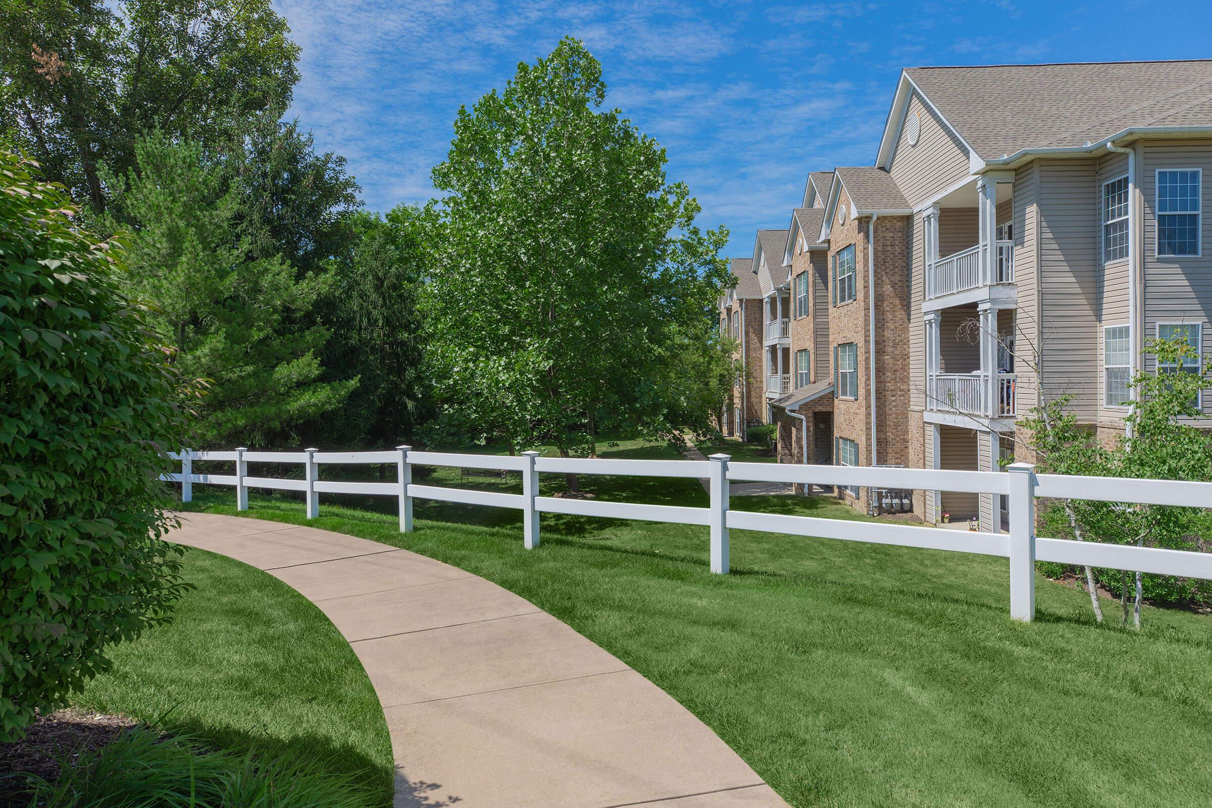 A landscaped path curves through a lush green lawn, flanked by a white fence. In the background, there are multi-story residential buildings with balconies, surrounded by trees under a bright blue sky with scattered clouds.