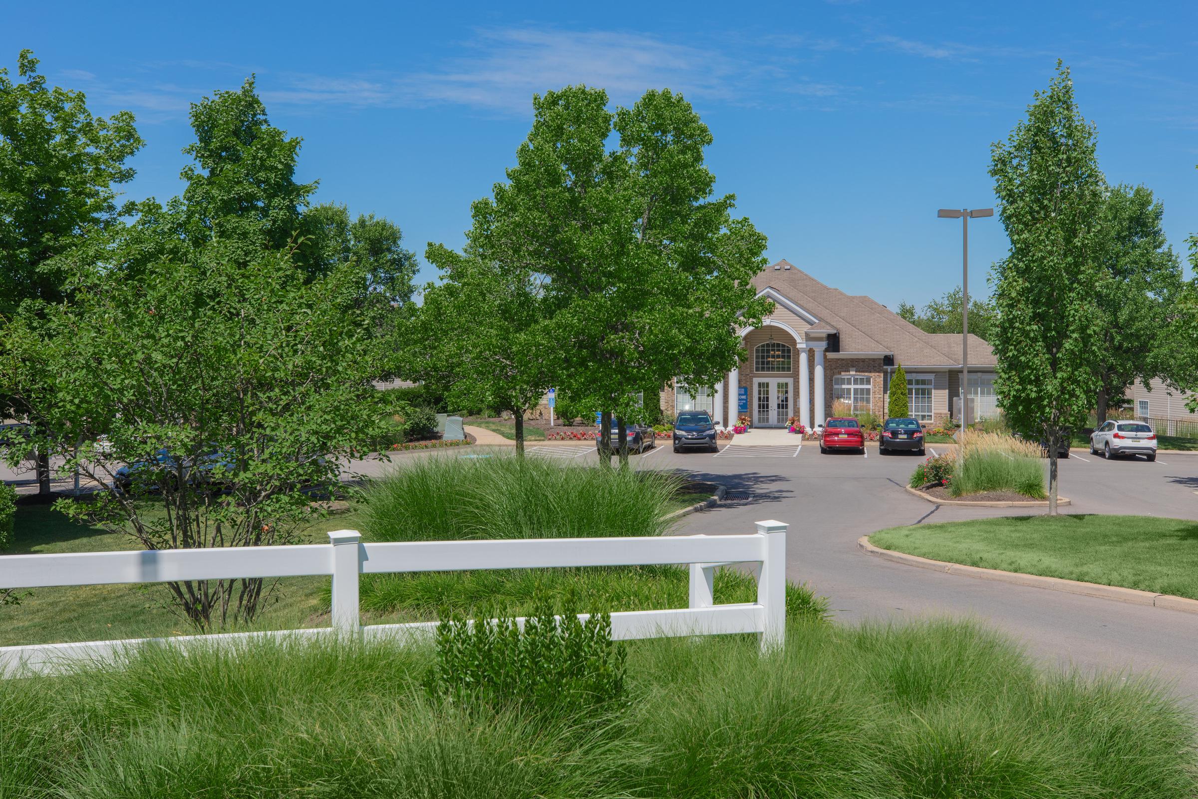 A landscaped view of a residential community entrance featuring a large building in the background, surrounded by green trees and well-maintained grass. Several cars are parked in the lot, under a clear blue sky.