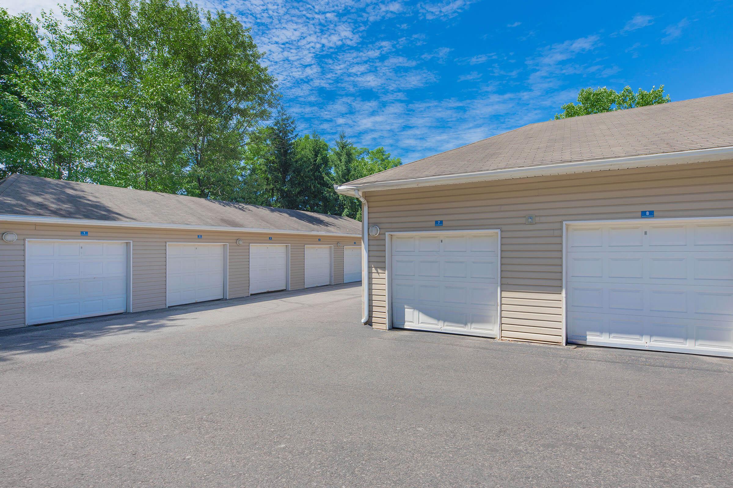 A row of garage doors in a light-colored building, set against a bright blue sky with scattered clouds and green trees in the background. The garages are well-maintained, with a paved driveway leading up to them.