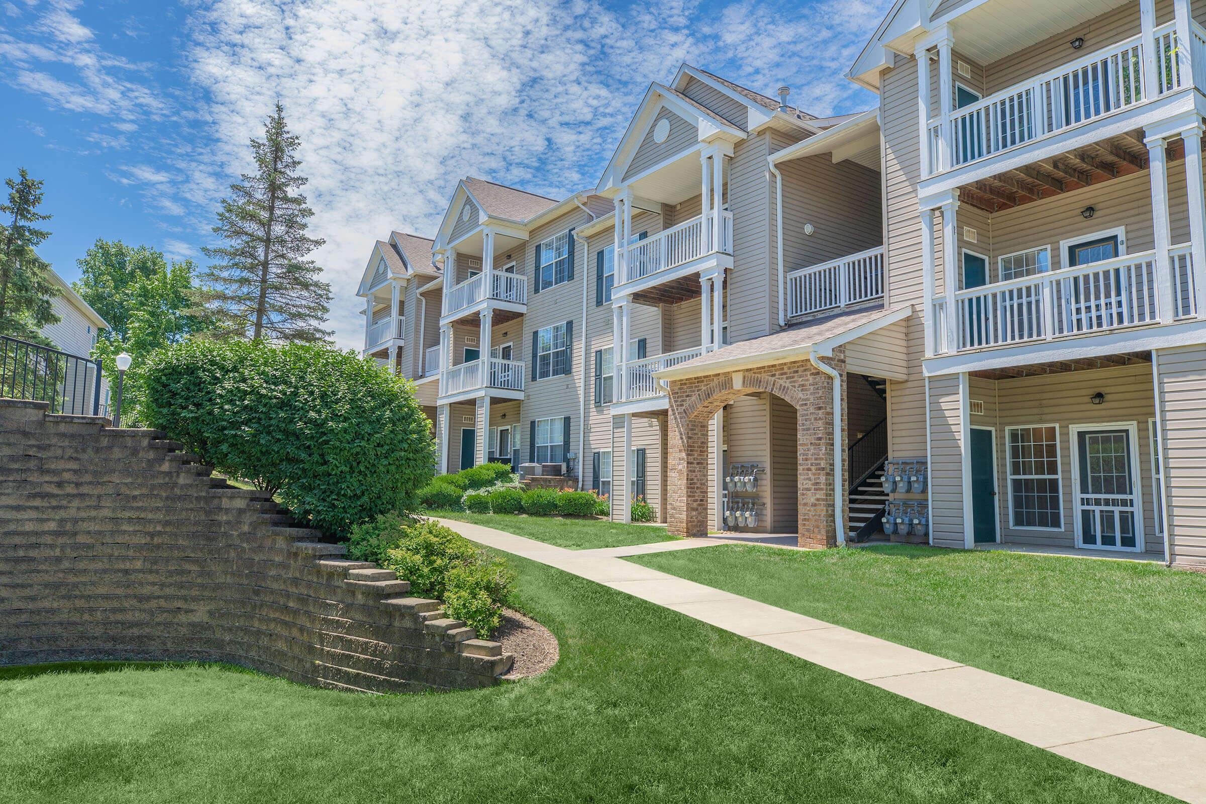 A well-maintained apartment community with multiple stories, surrounded by lush green grass and landscaped areas. The buildings feature balconies and patio spaces, and a paved walkway leads through the property, with a stone retaining wall beside it.