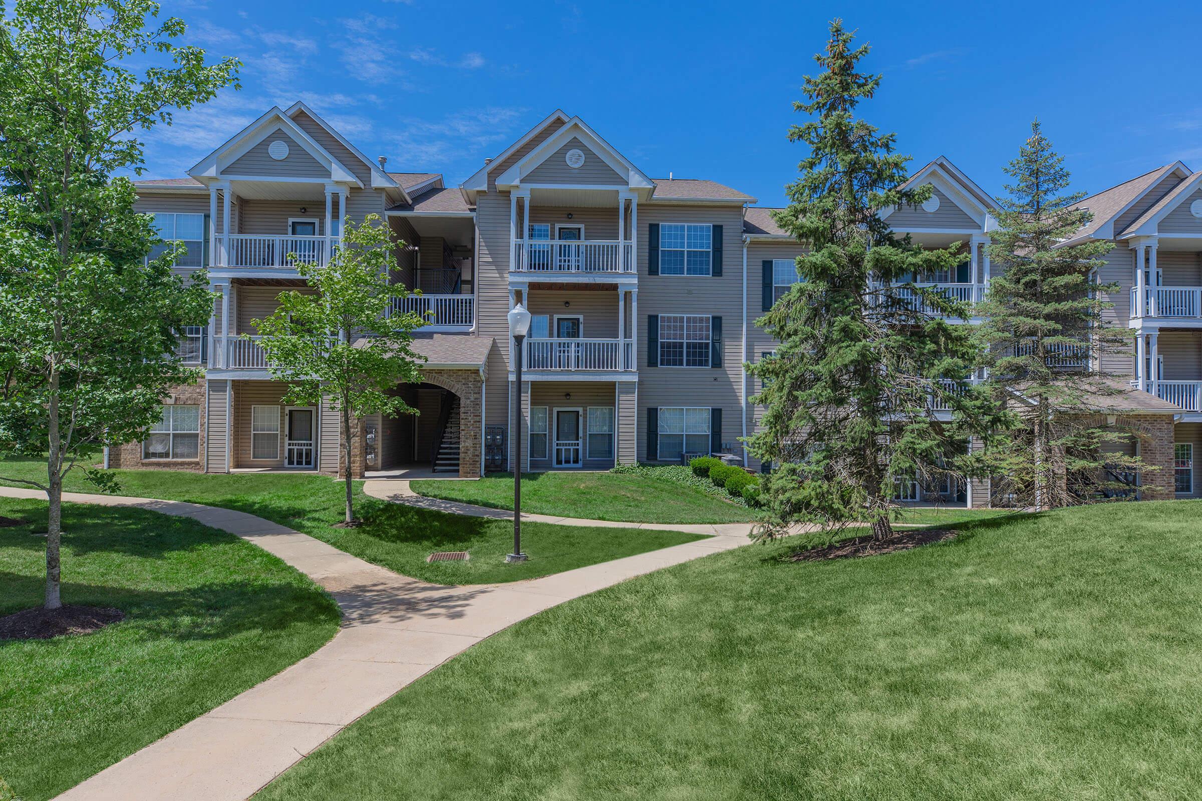 A well-maintained apartment community with multiple balconies, surrounded by green lawns and trees, under a clear blue sky. Pathways lead through the landscaped area.