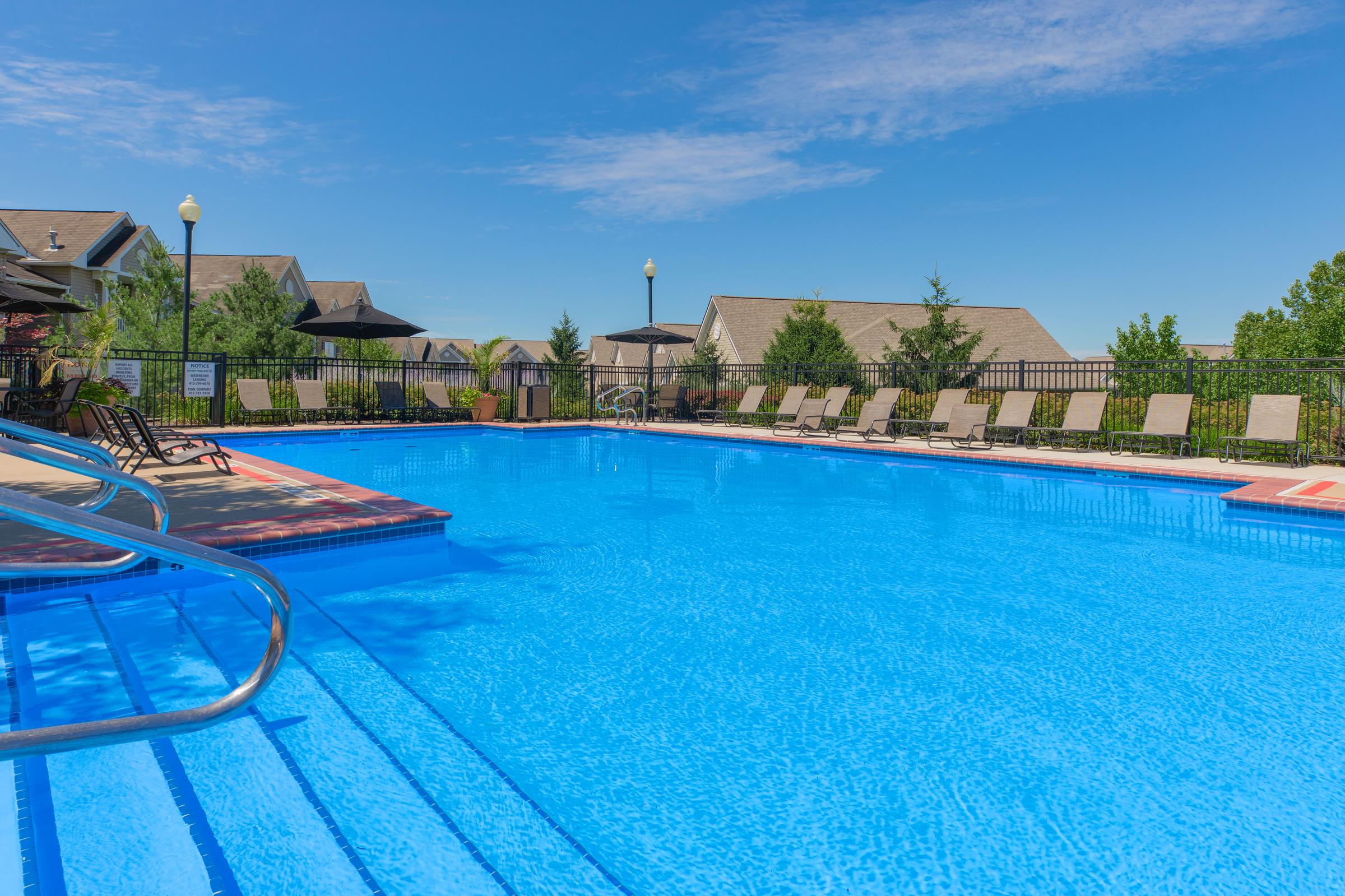 A clear blue swimming pool surrounded by lounge chairs and umbrellas, with residential buildings and trees in the background under a bright blue sky.