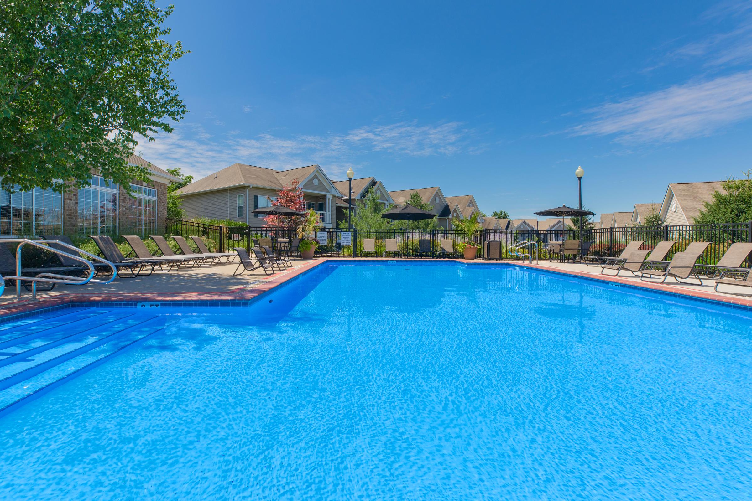 A clear blue swimming pool surrounded by lounge chairs, with residential buildings in the background and a bright blue sky above.