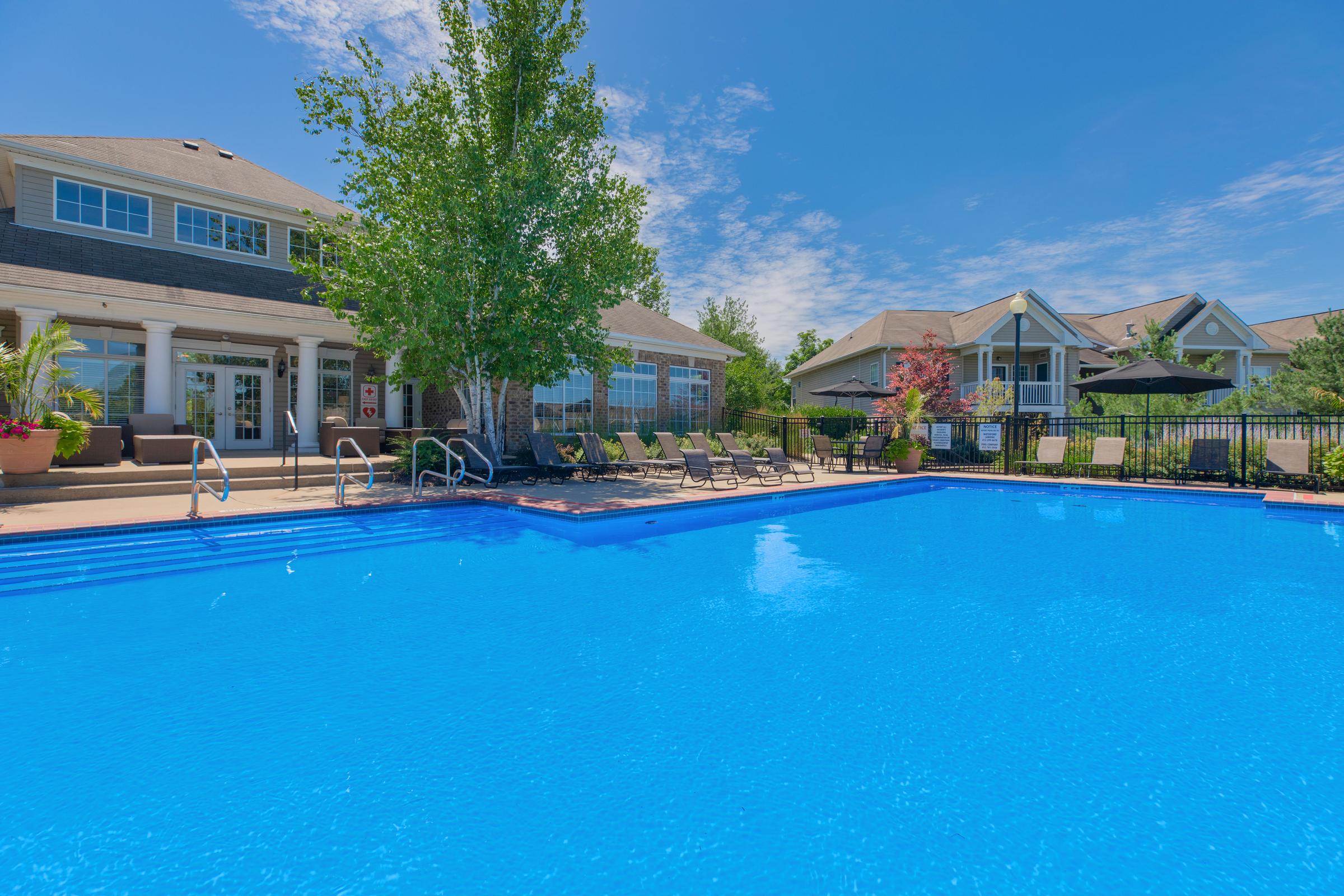 A bright blue swimming pool in a residential area, surrounded by lounge chairs and lush greenery. In the background, there are well-maintained buildings with porches and umbrellas providing shade, under a clear blue sky.