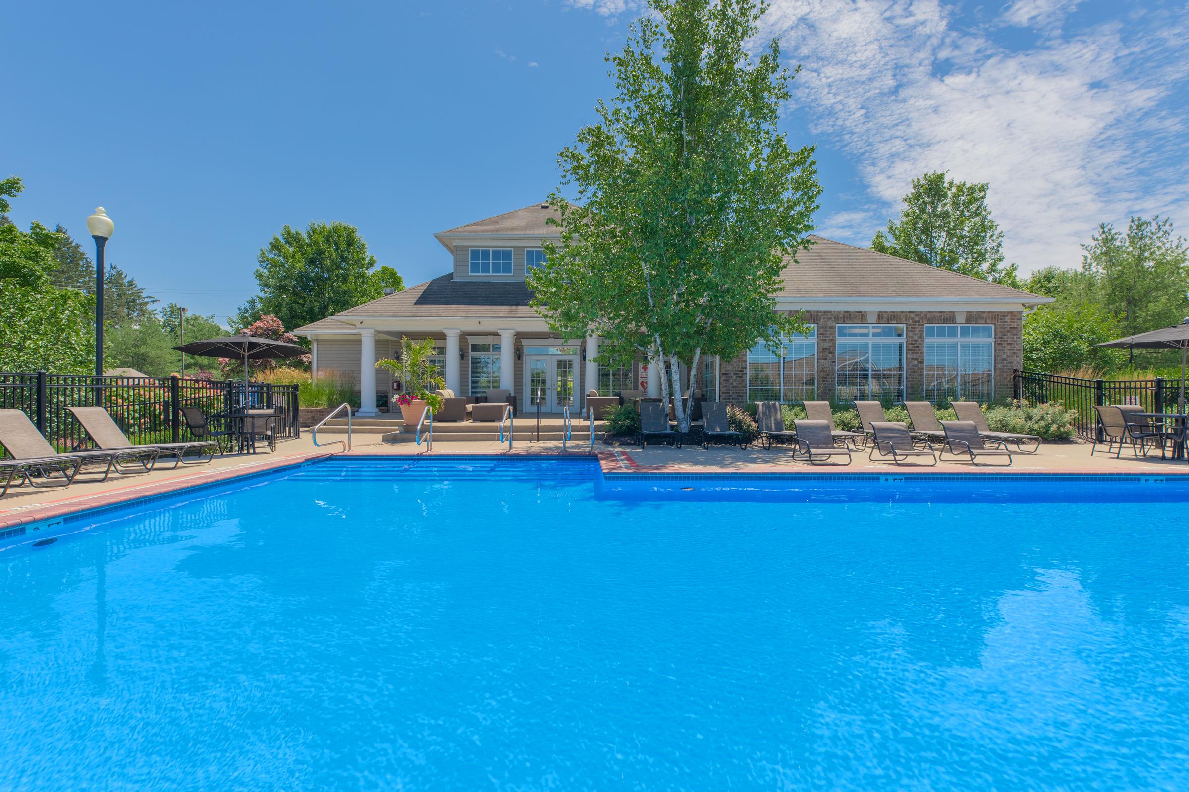 A clear blue swimming pool surrounded by lounge chairs and shaded by umbrellas, with a residential building in the background and trees adding greenery to the scene. The sky is bright and partly cloudy.