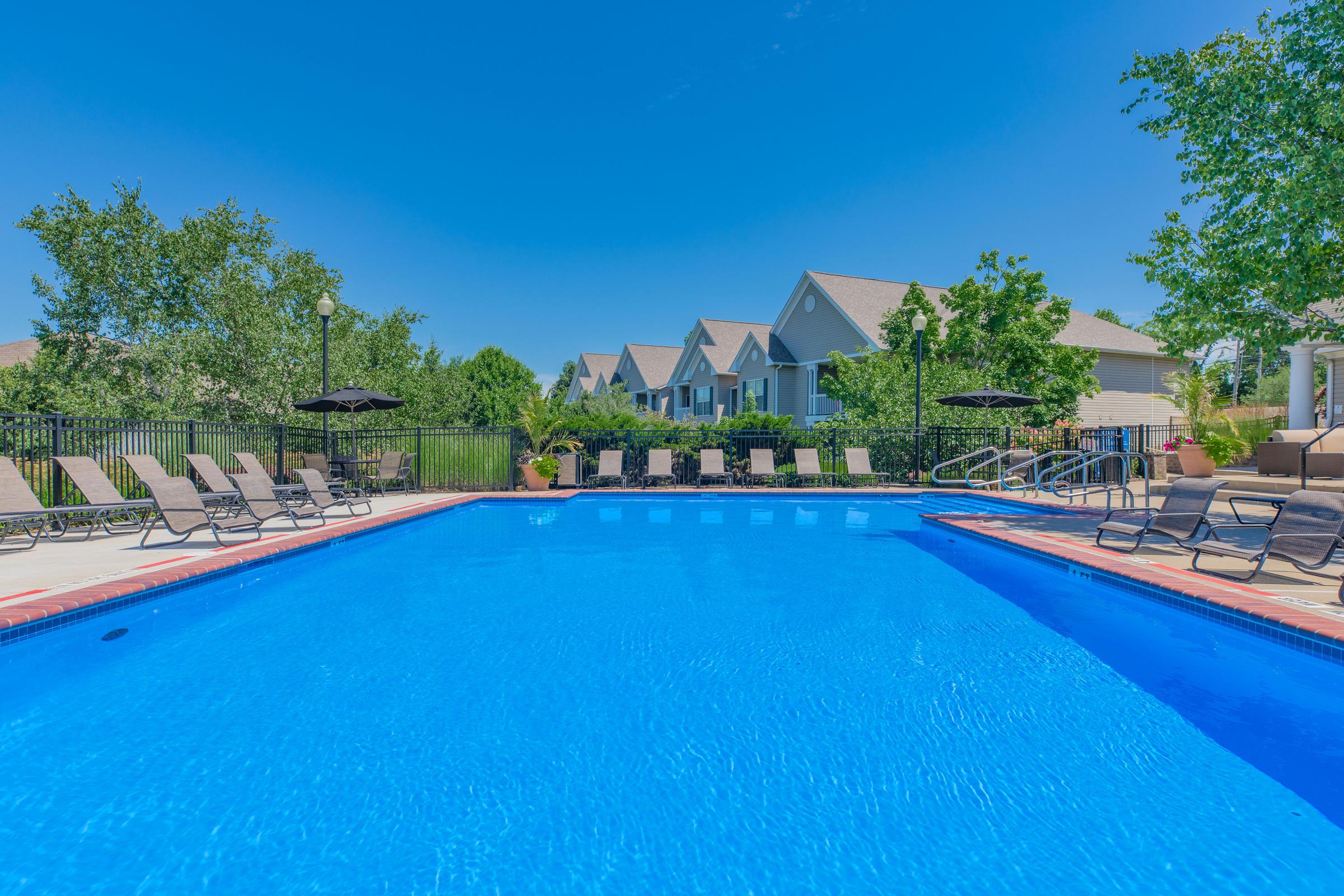 A swimming pool with clear blue water surrounded by lounge chairs and umbrellas, set against a backdrop of green trees and residential buildings under a clear blue sky.