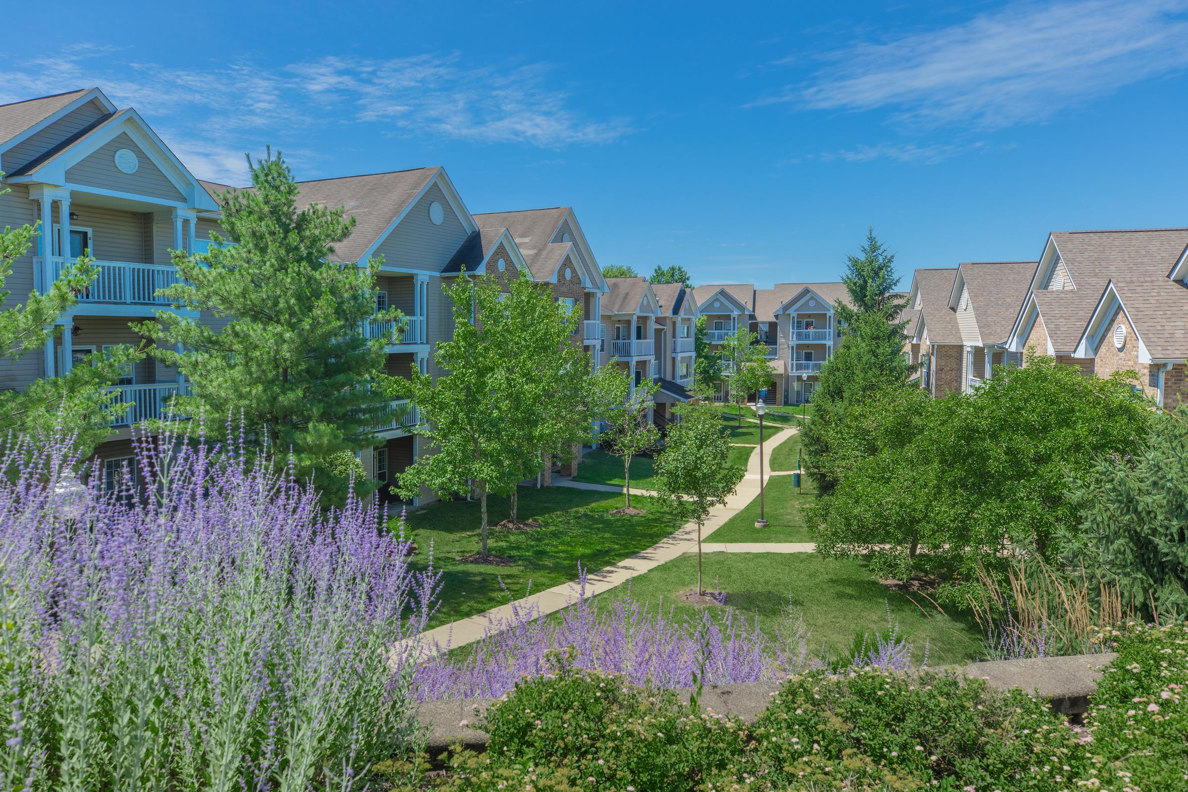 A sunny view of a residential apartment community featuring well-maintained buildings, green lawns, and blooming purple flowers. Pathways weave through lush landscaping, with trees providing shade in a peaceful environment.