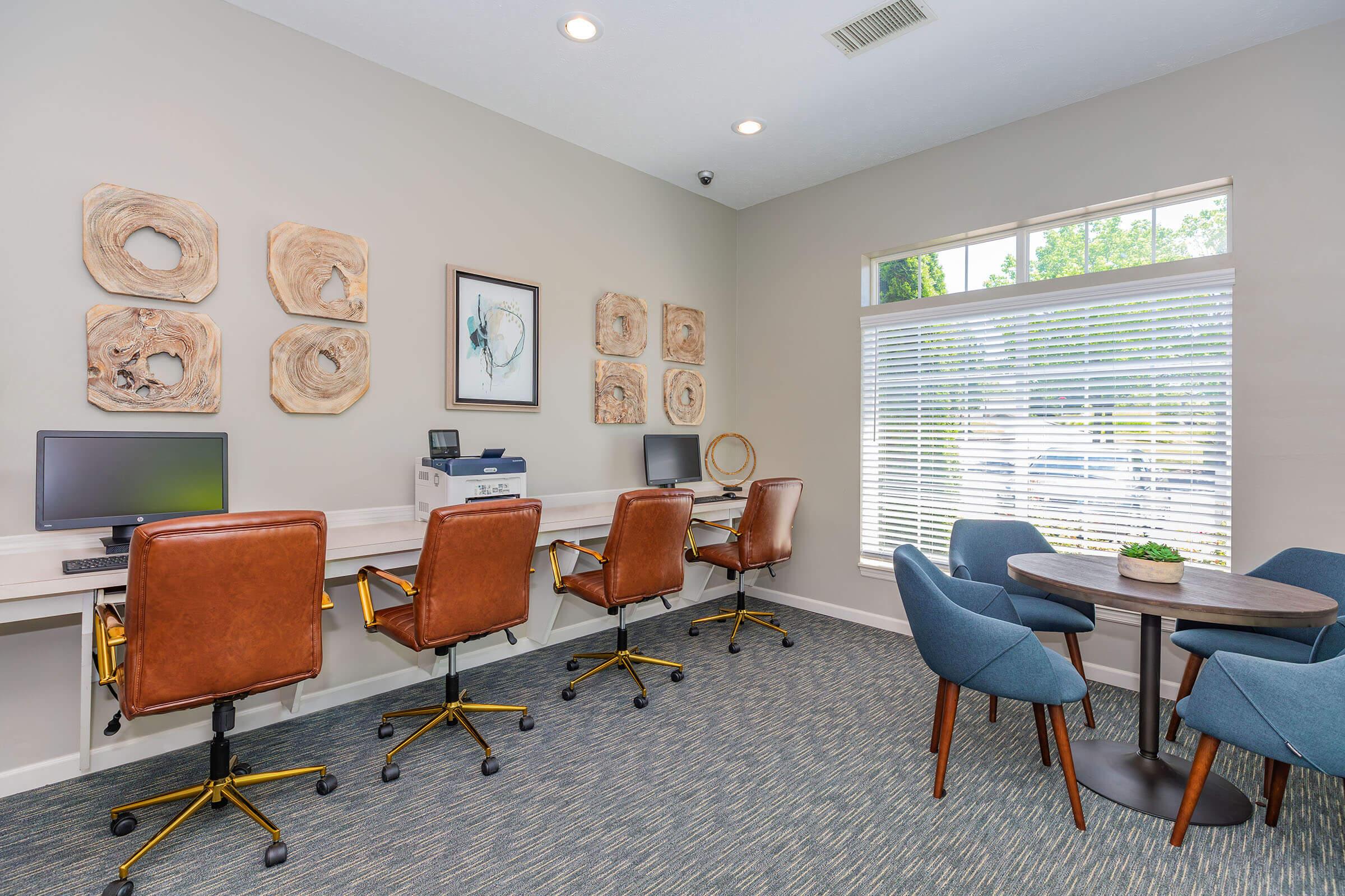 A modern office space featuring four brown leather office chairs at a white desk with two desktop computers. A printer is also on the desk. The walls are adorned with circular wooden artwork, and there is a small round table with blue chairs near a window with blinds letting in natural light.