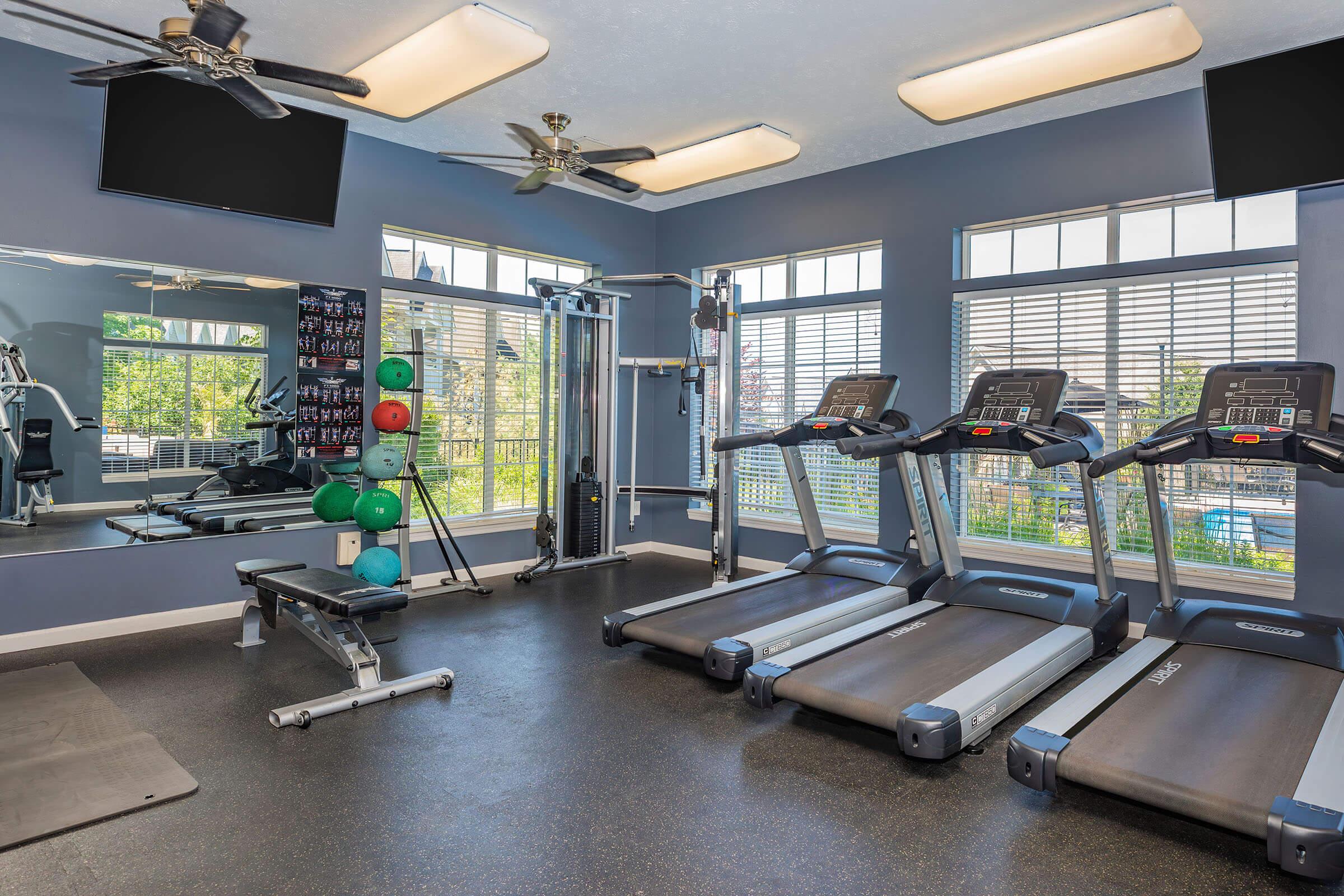 A modern gym interior featuring two treadmills, a weight bench, various weights, and exercise equipment. Large windows provide natural light, and flat-screen TVs are mounted on the walls.