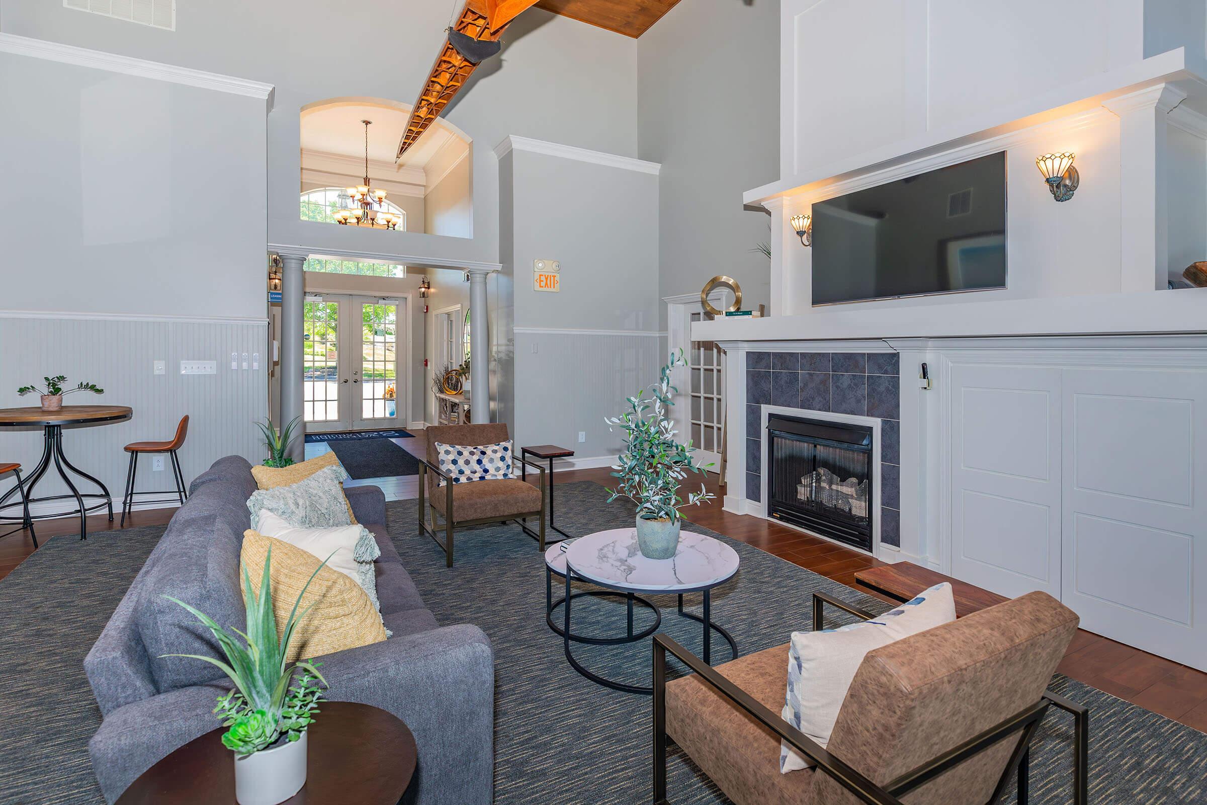 A spacious living room featuring a comfortable gray sofa with decorative cushions, two modern chairs, and a round coffee table with a plant. The room has a large fireplace with a flat-screen TV above it, high ceilings with wooden beams, and large windows allowing natural light to fill the space. Decorative plants and stylish furnishings add to the inviting atmosphere.