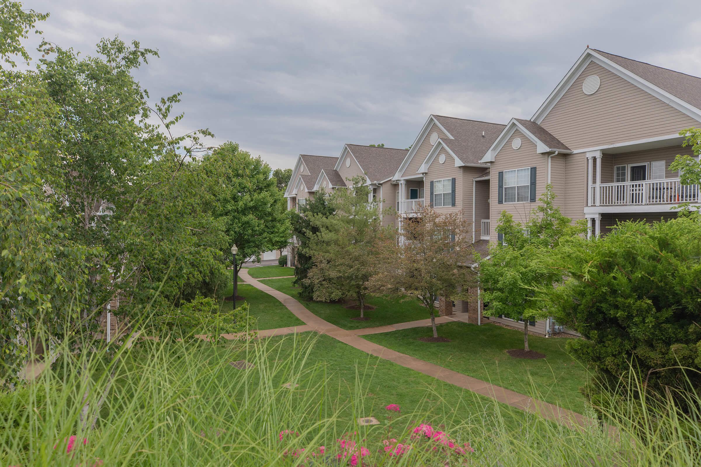 A scenic view of a residential area featuring multiple buildings arranged along pathways, surrounded by greenery and trees, under a cloudy sky.