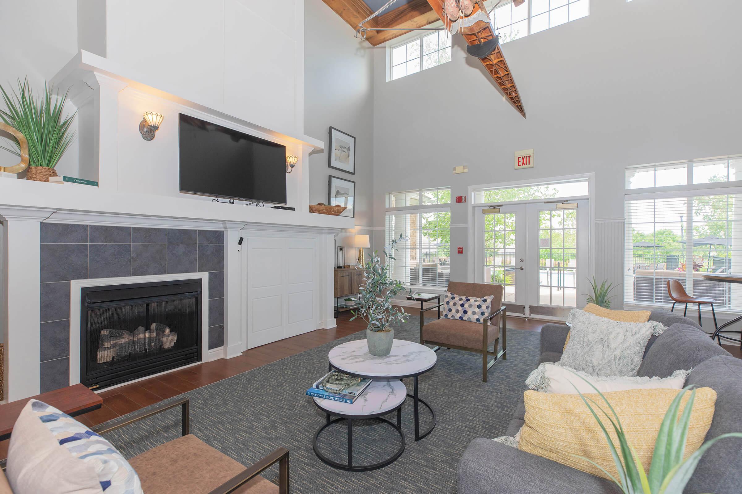 Bright and modern living room with high ceilings, featuring a cozy fireplace, large windows, and stylish furnishings. A comfortable sofa with decorative pillows is paired with a circular coffee table, while a large TV is mounted above the fireplace. There are plants and artwork adding a homey touch.