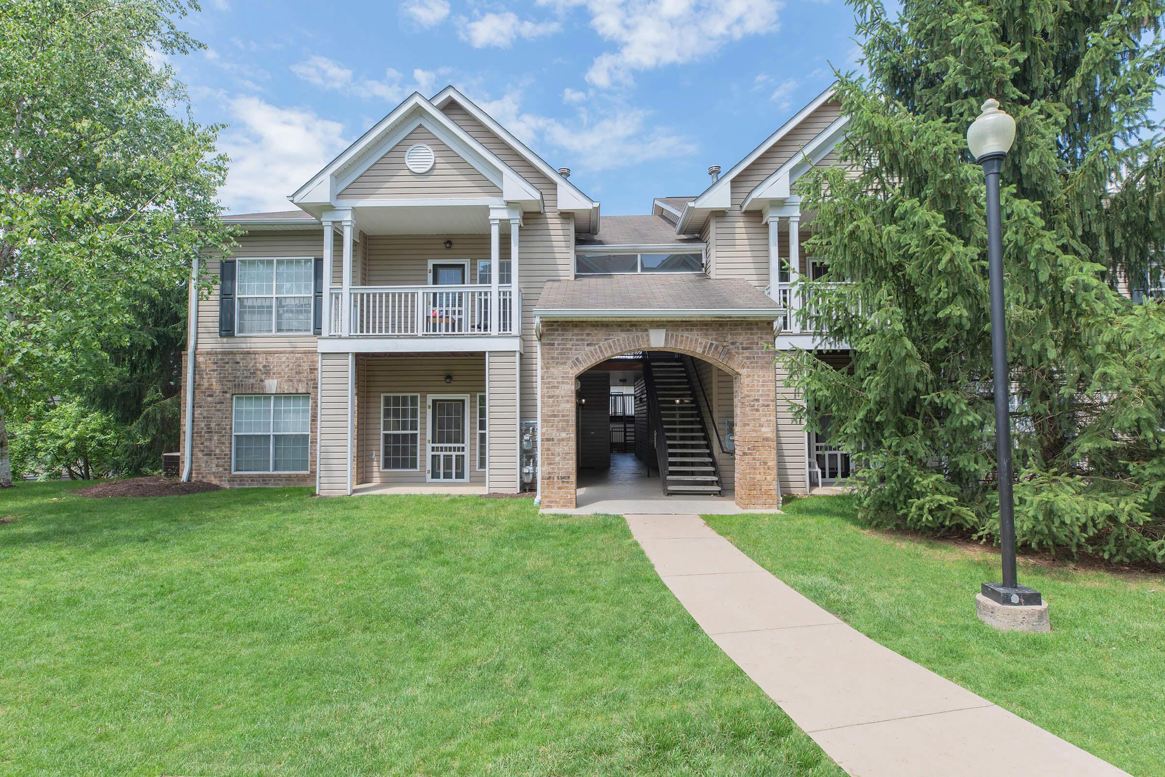 A two-story apartment building with a landscaped lawn, featuring balconies and an entrance with decorative stonework. Green trees are visible on either side of the building, and a pathway leads to the entrance.