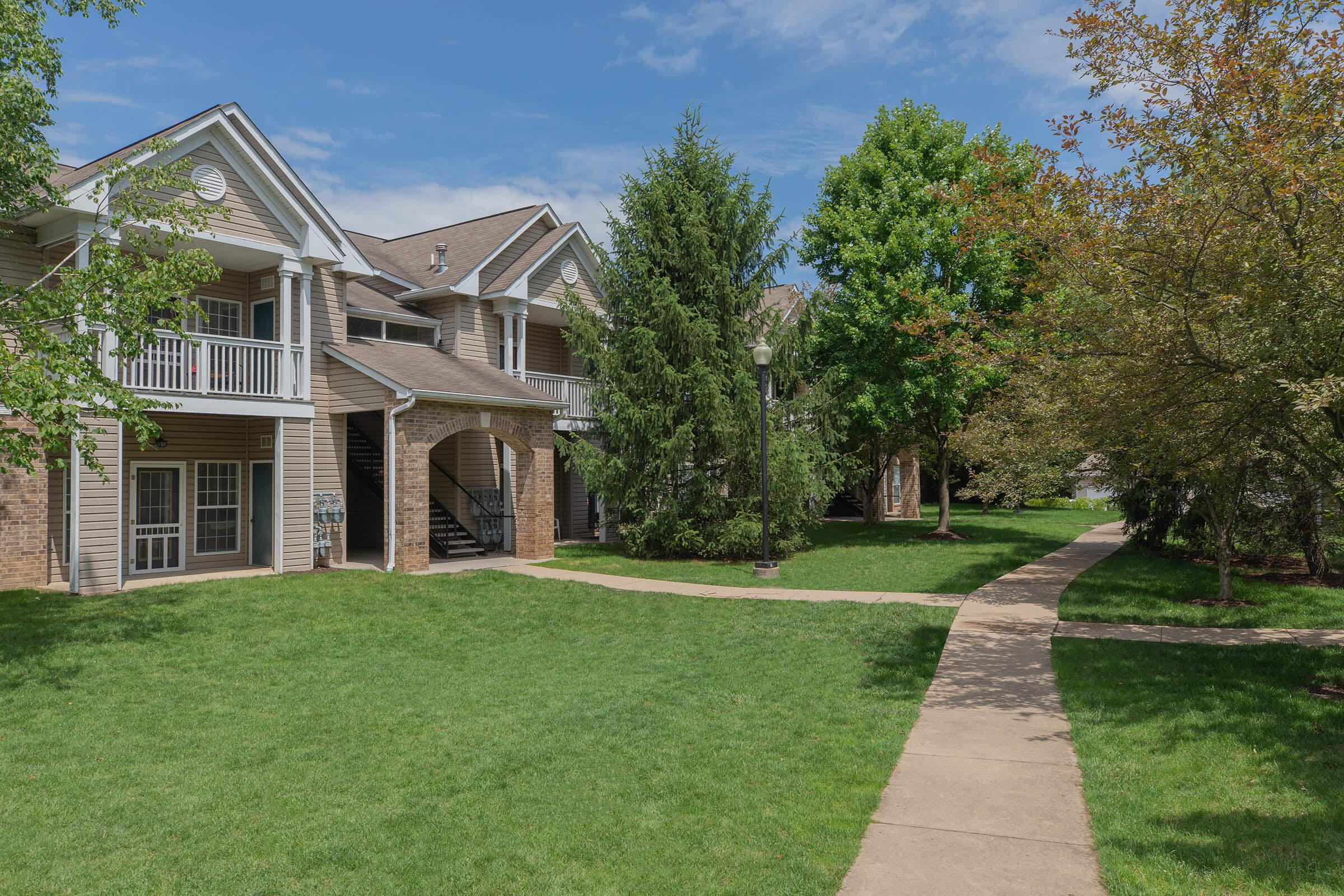 A landscaped area featuring a two-story apartment building with balconies, surrounded by grass and trees. A concrete pathway winds through the greenery, leading to additional areas. The sky is partly cloudy.