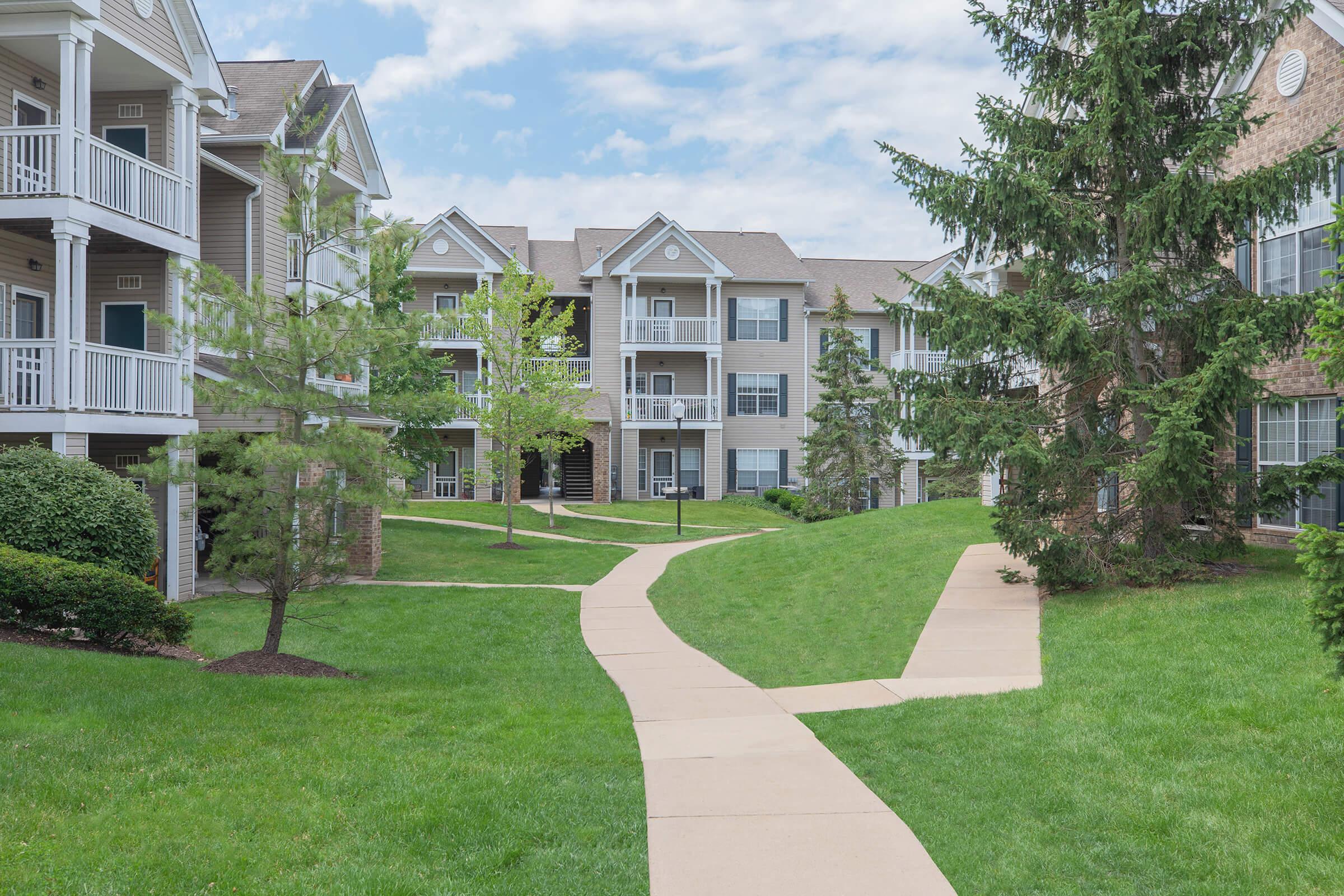 A well-maintained residential complex featuring multiple buildings with balconies, surrounded by landscaped green lawns and trees. A concrete pathway winds through the grassy area, providing access to the entrances of the buildings. The sky is partly cloudy.