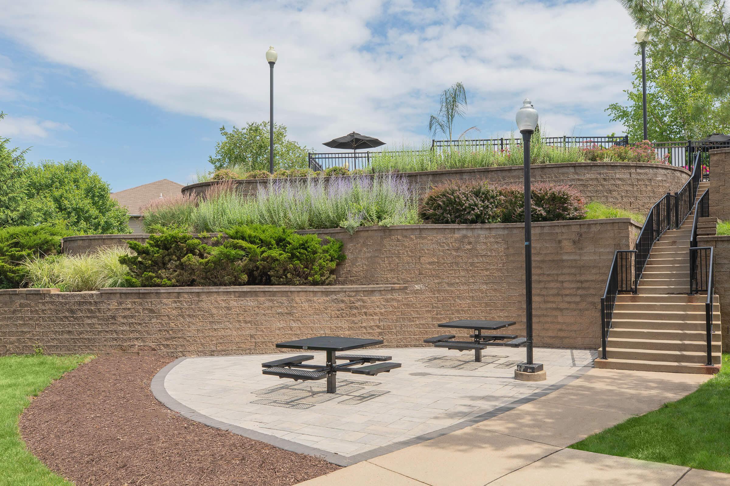 A landscaped outdoor area featuring stone pathways, three picnic tables, and decorative plants on an elevated section with a staircase leading up. There are street lamps and a shaded umbrella at the top surrounded by greenery.