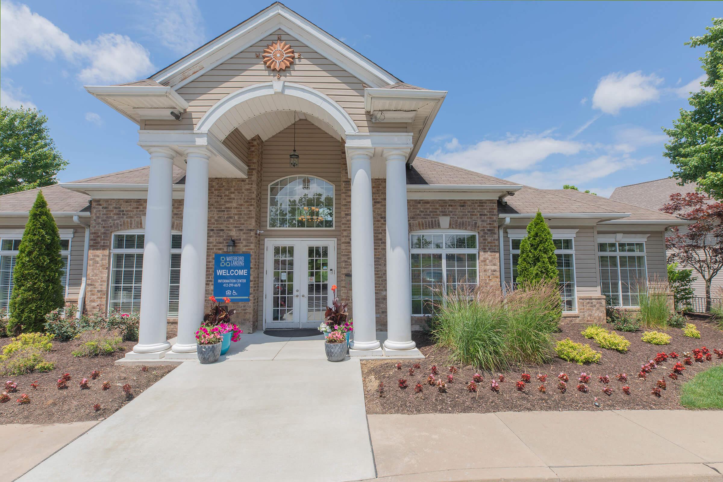 A welcoming entrance of a residential building featuring a large front porch with columns, decorative landscaping, and potted plants. A sign indicating "Welcome" is prominently displayed. Clear blue skies and well-manicured bushes enhance the inviting atmosphere.