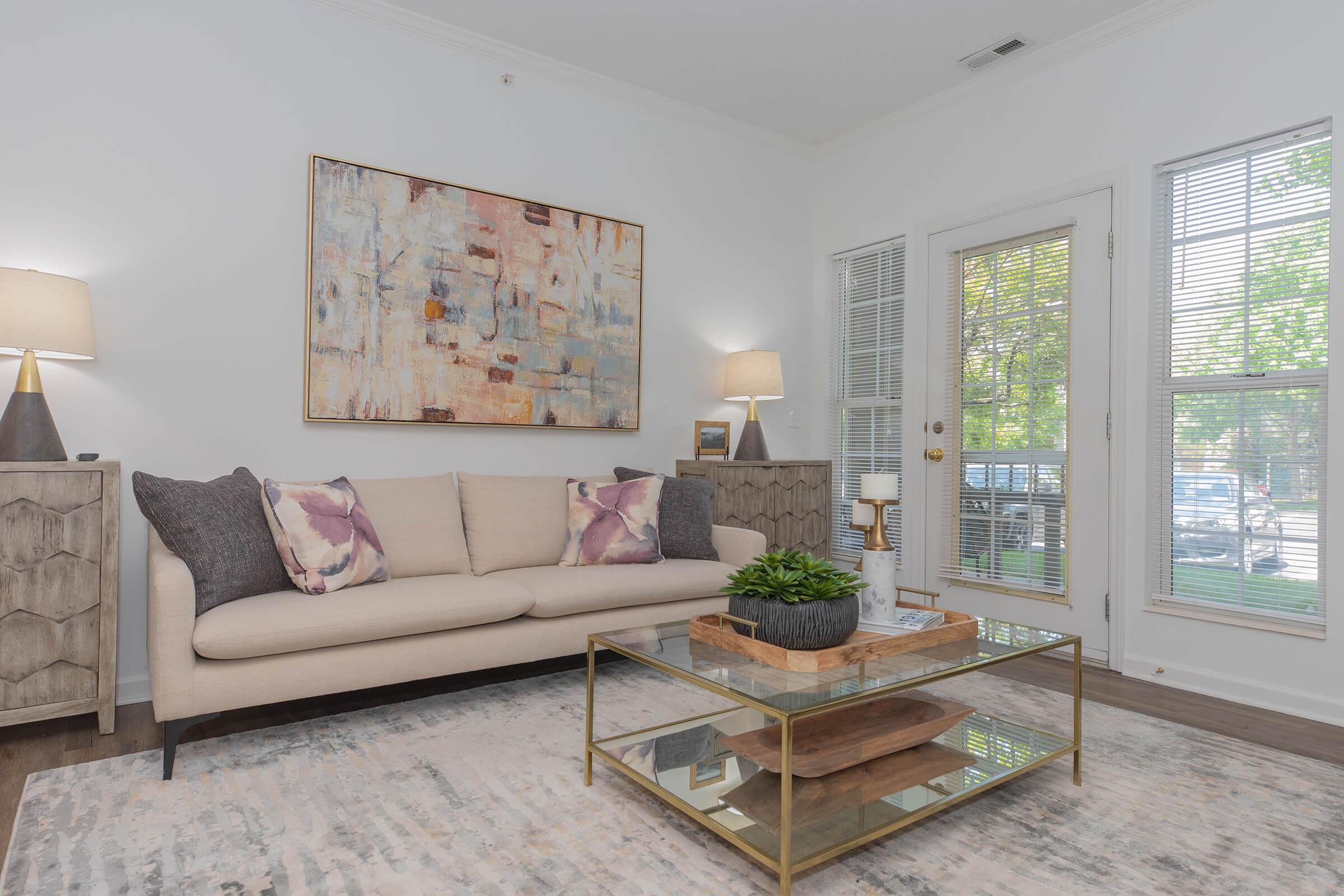 A modern living room featuring a light-colored sofa adorned with decorative pillows, a glass coffee table with a plant centerpiece, a textured area rug, and framed abstract wall art. The room is bright with natural light coming through a nearby window, flanked by lamps on either side.