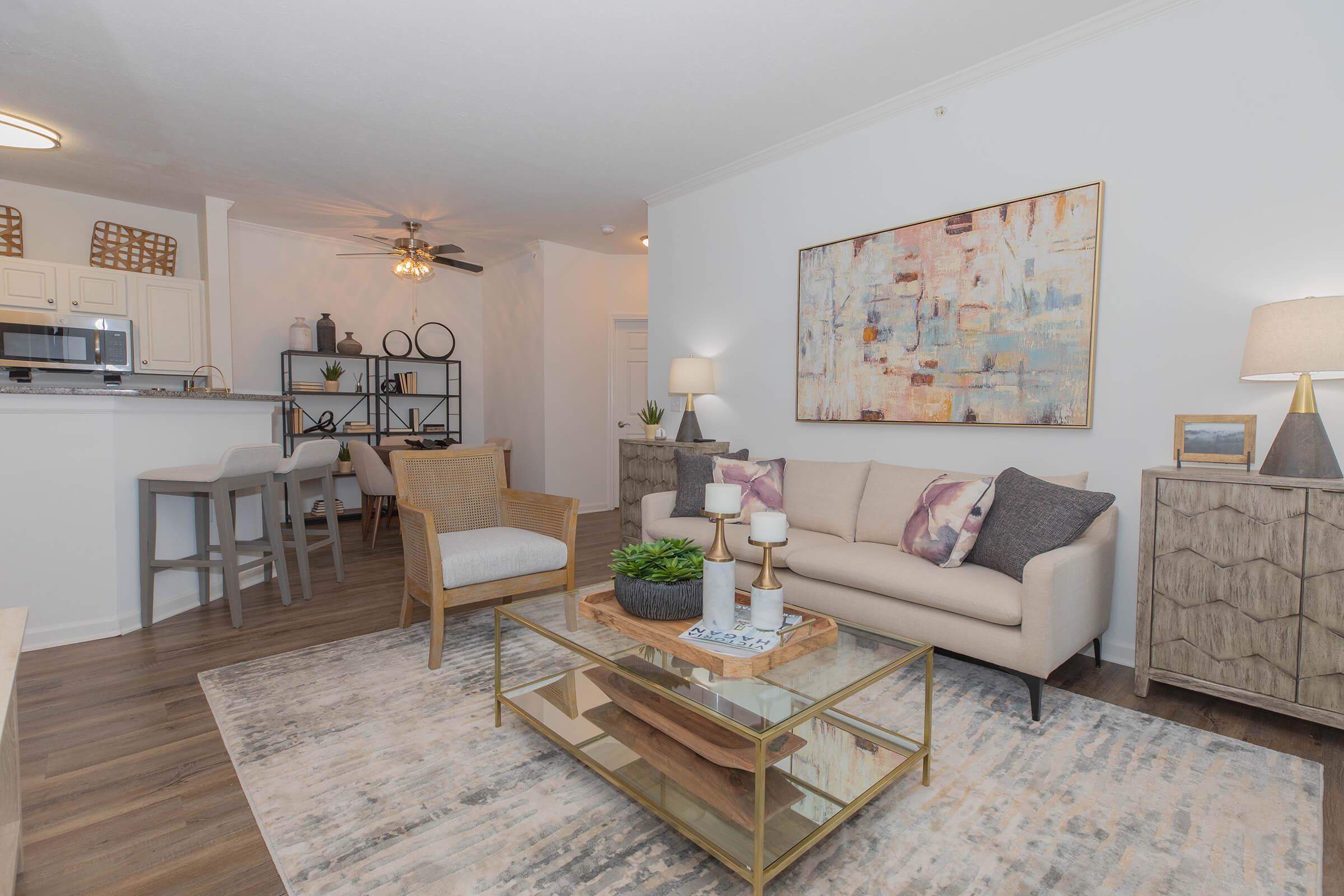 A modern living room featuring a light-colored sofa with decorative pillows, a glass coffee table, and stylish accessories. The space includes an area rug, a shelf with decorative items, and a kitchenette in the background. Warm lighting creates a cozy atmosphere.