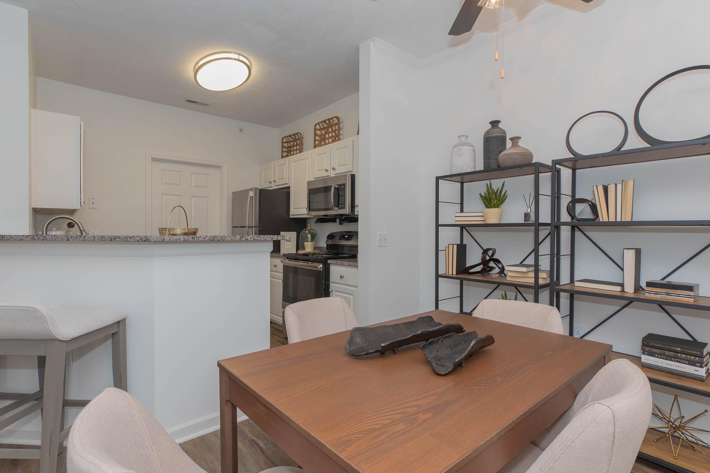 A modern dining area featuring a wooden table with decorative objects, surrounded by light-colored chairs. In the background, a well-equipped kitchen with stainless steel appliances and white cabinetry is visible. A stylish bookshelf displays various decorative items and a small plant.