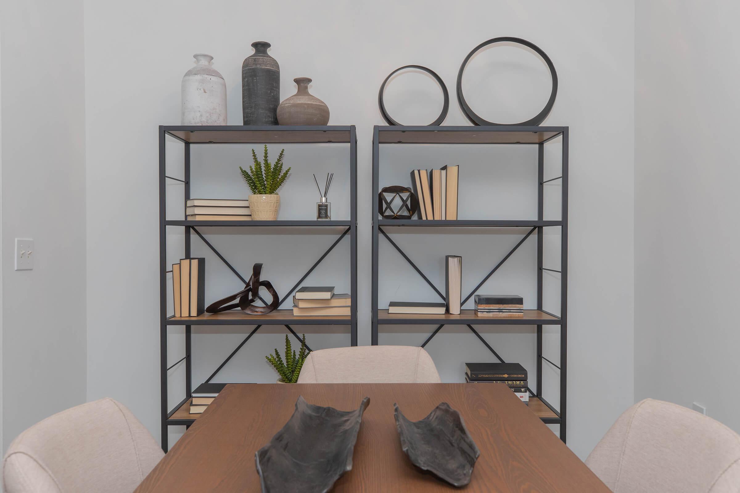 A modern interior scene featuring two black metal bookshelves filled with books, decorative objects, and plants. The shelves are arranged against a light-colored wall. In the foreground, a wooden dining table is set with two abstract black decorative pieces, and light beige chairs are positioned around it.