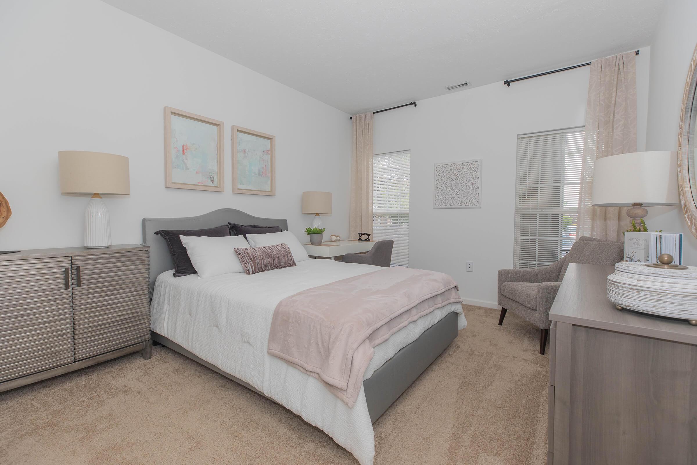 A modern, stylish bedroom featuring a queen-sized bed with white bedding and decorative pillows. Two framed artworks hang above the bed. A nightstand with a lamp is on one side of the bed, while a gray armchair and a dresser with decorative items are on the opposite side. Light-colored curtains frame the windows, creating a cozy atmosphere.