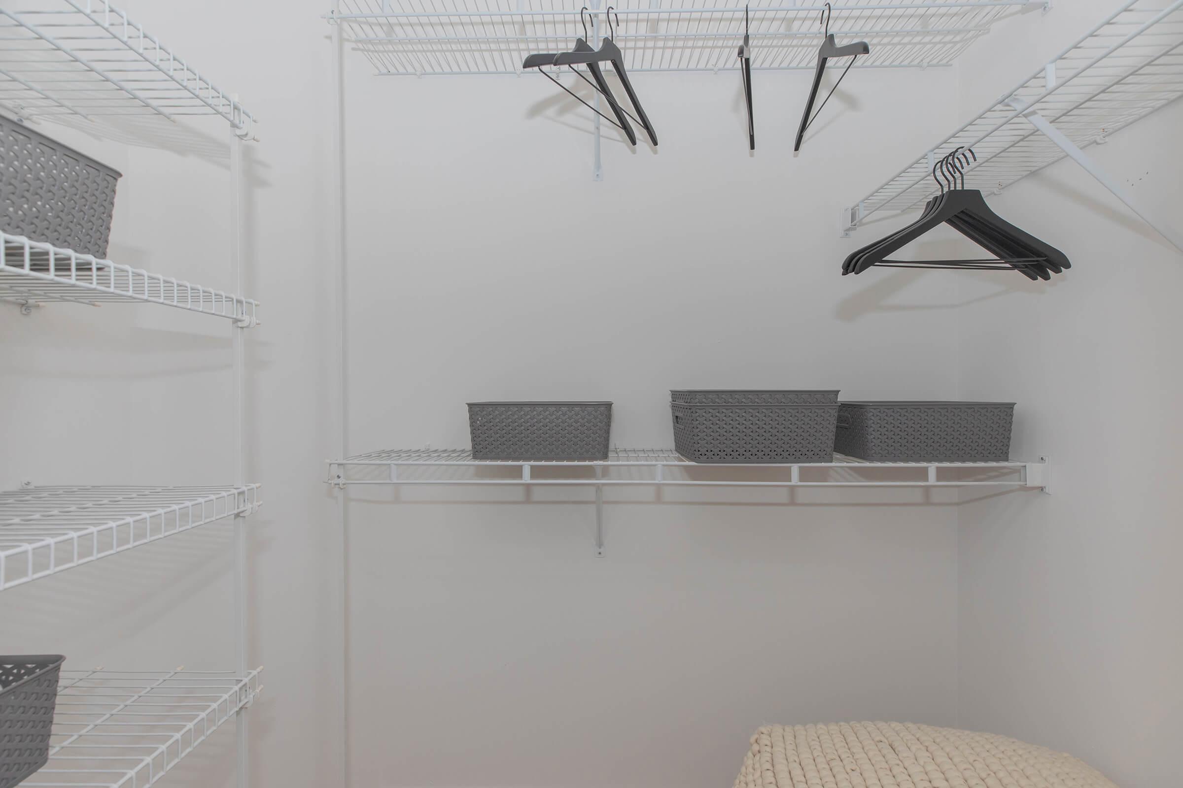 A clean, empty closet with white wire shelving. There are a few gray baskets on one shelf and several black hangers hanging from another shelf. The closet has a neutral white background, creating a spacious and organized appearance.