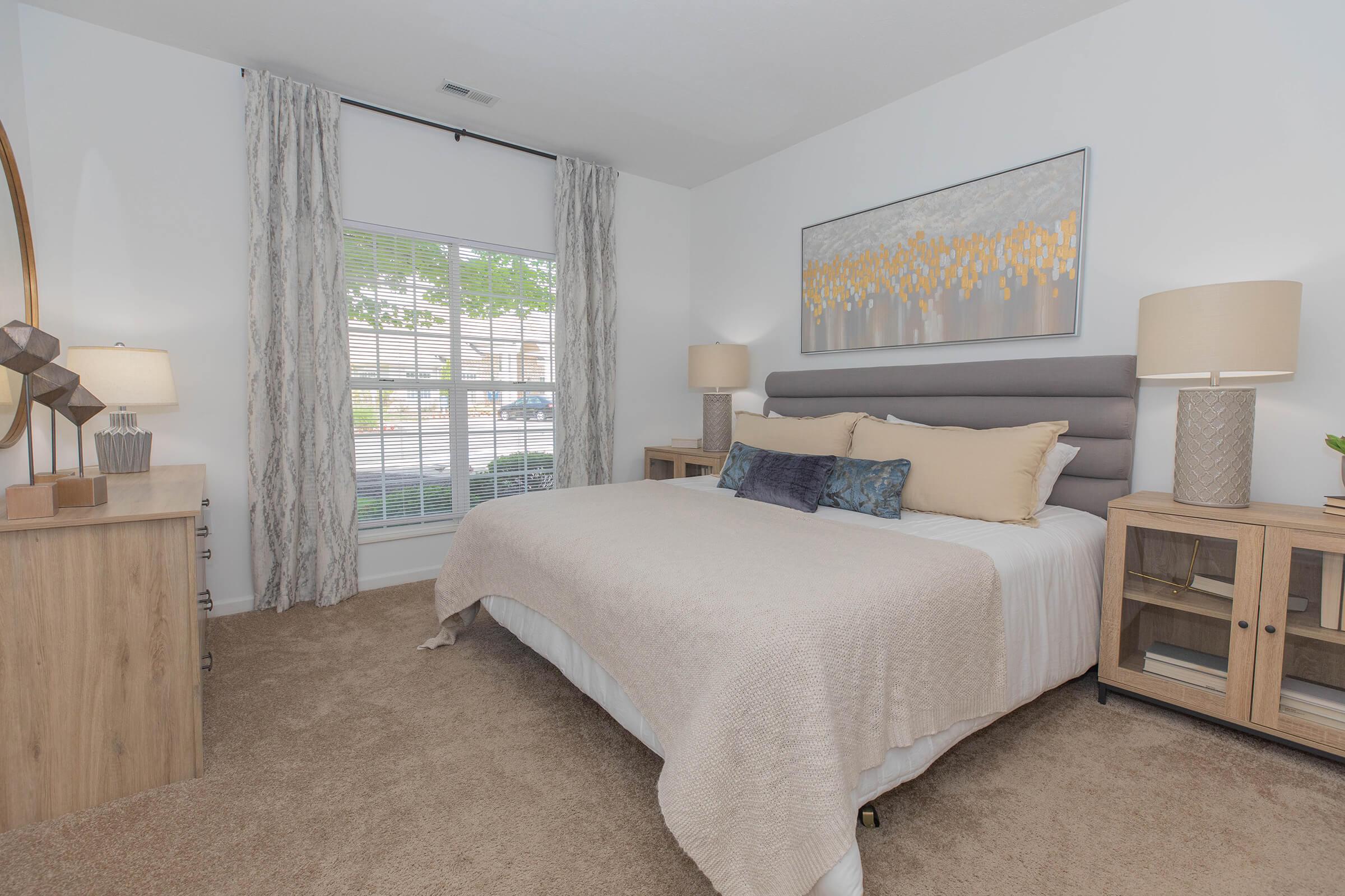 A cozy bedroom featuring a neatly made bed with a light beige comforter and decorative pillows. On either side of the bed are wooden nightstands with lamps. A large window with light curtains lets in natural light, and a piece of artwork hangs above the bed, adding a touch of color to the room.