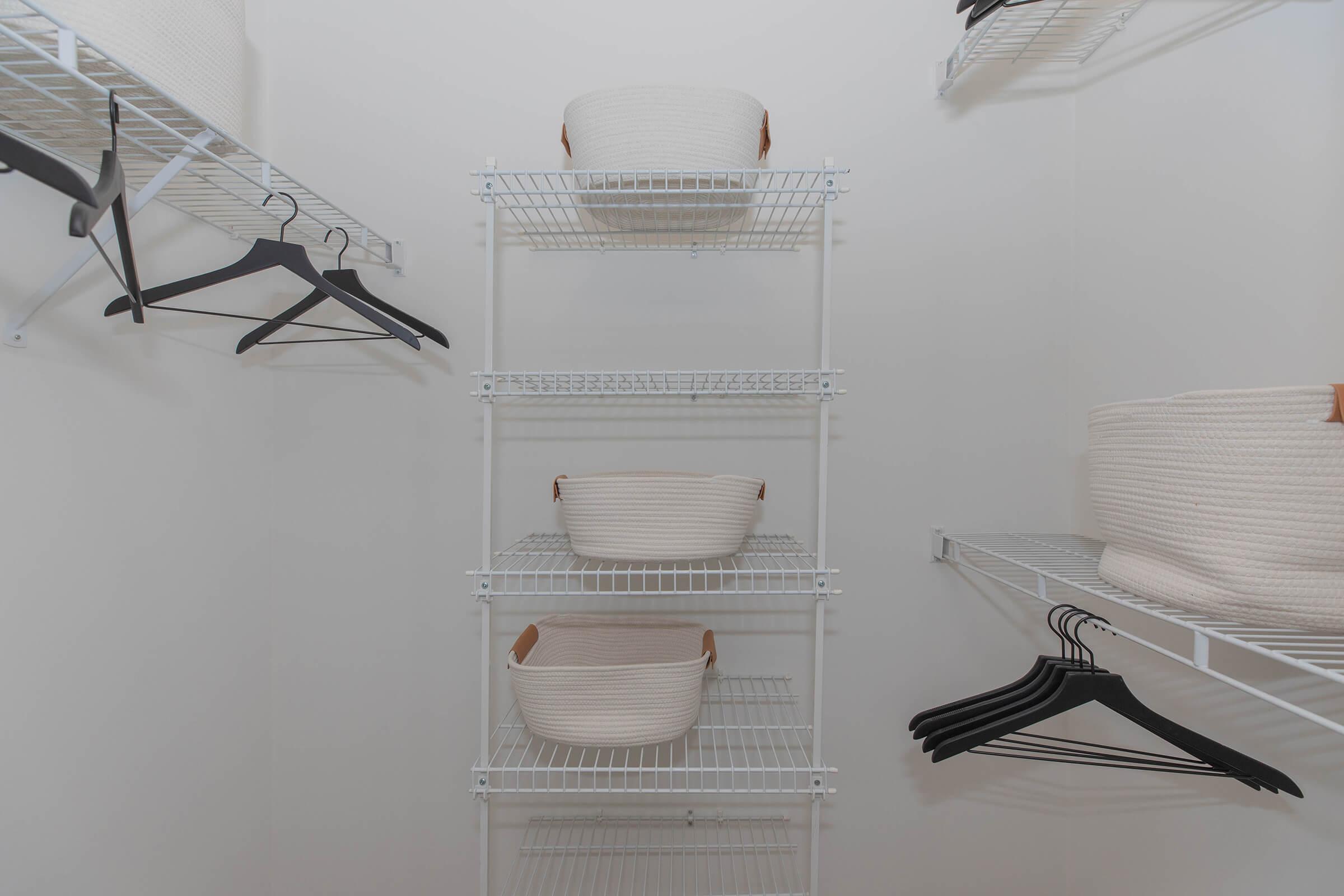 A clean, organized closet with white wire shelving. The shelves hold woven baskets and several black hangers hang on the side. The background is a plain white wall, emphasizing the tidy arrangement of storage items.