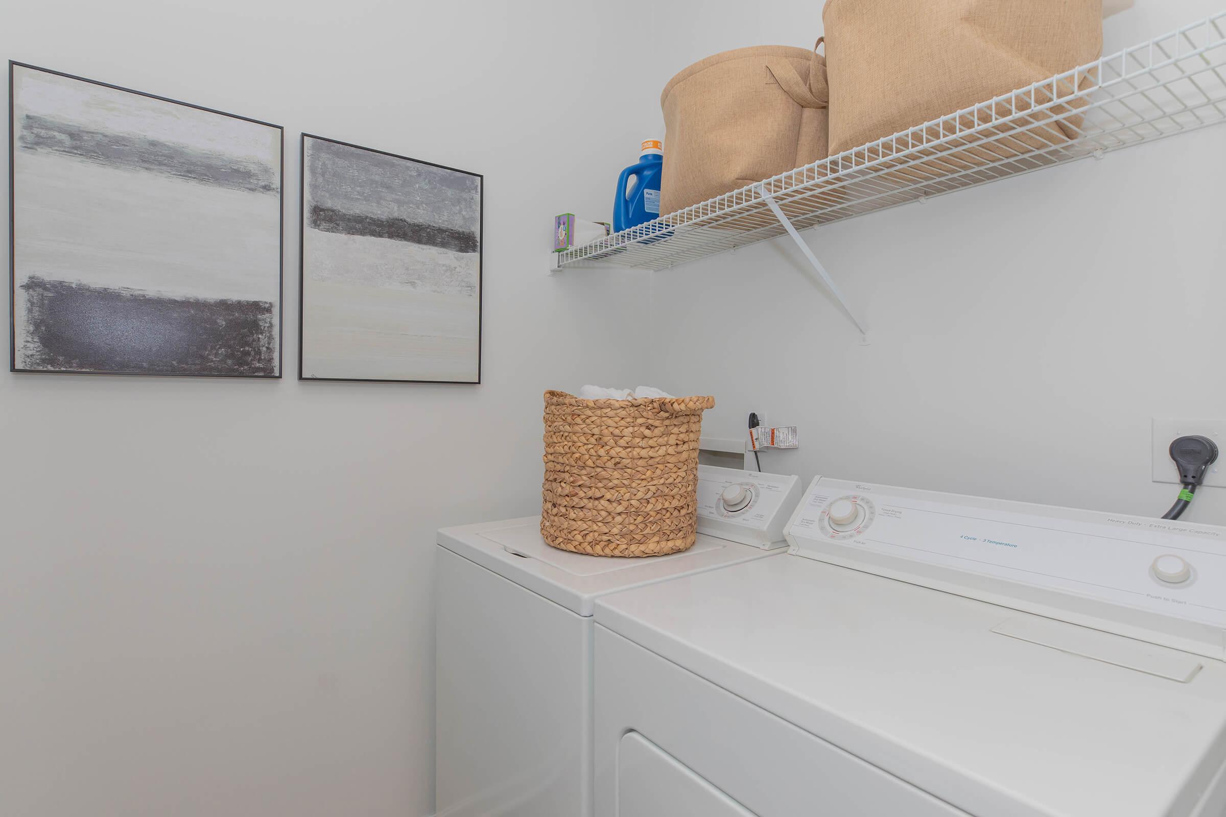 A bright laundry room featuring two abstract framed artworks on the wall, a white washing machine and dryer, a woven basket filled with laundry on top, and a shelf above holding detergent and other supplies. The overall design is clean and organized, with a neutral color palette.