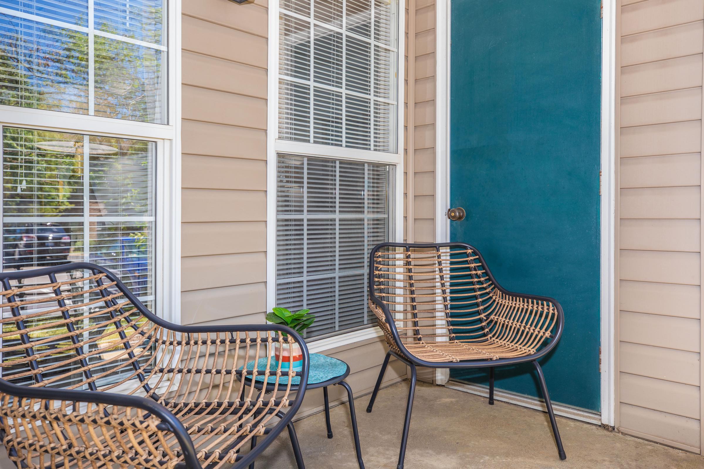 A cozy porch area featuring two wicker chairs and a small teal table. A bright, teal door is visible, along with large windows that allow natural light. The setting has a relaxed atmosphere, with a potted plant on the table and in a well-maintained outdoor space.