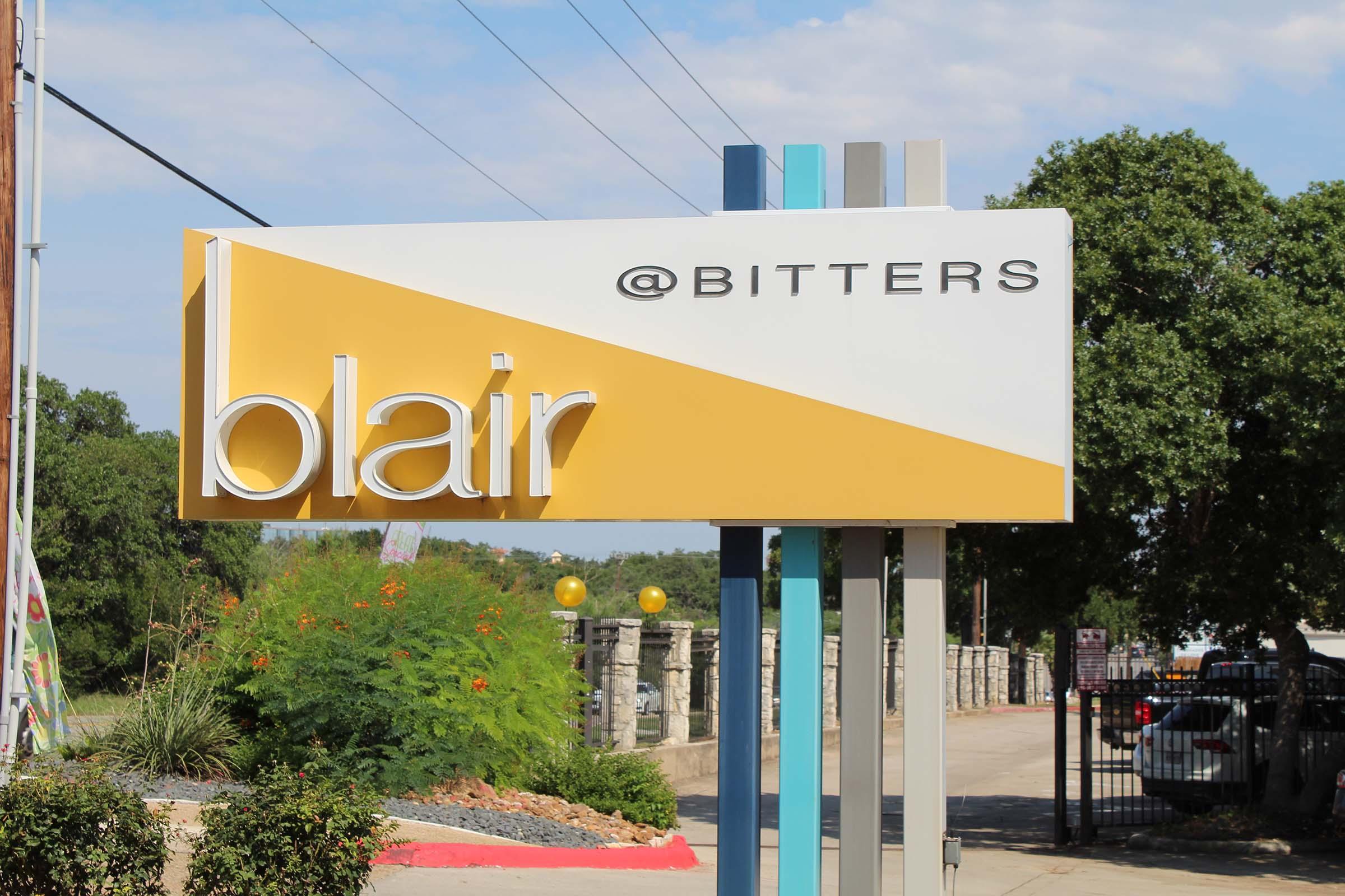 A sign featuring the word "blair" in bold white letters on a yellow and white background, with the text "@BITTERS" above it. The sign is positioned in an area with greenery and is visible on a clear, sunny day.