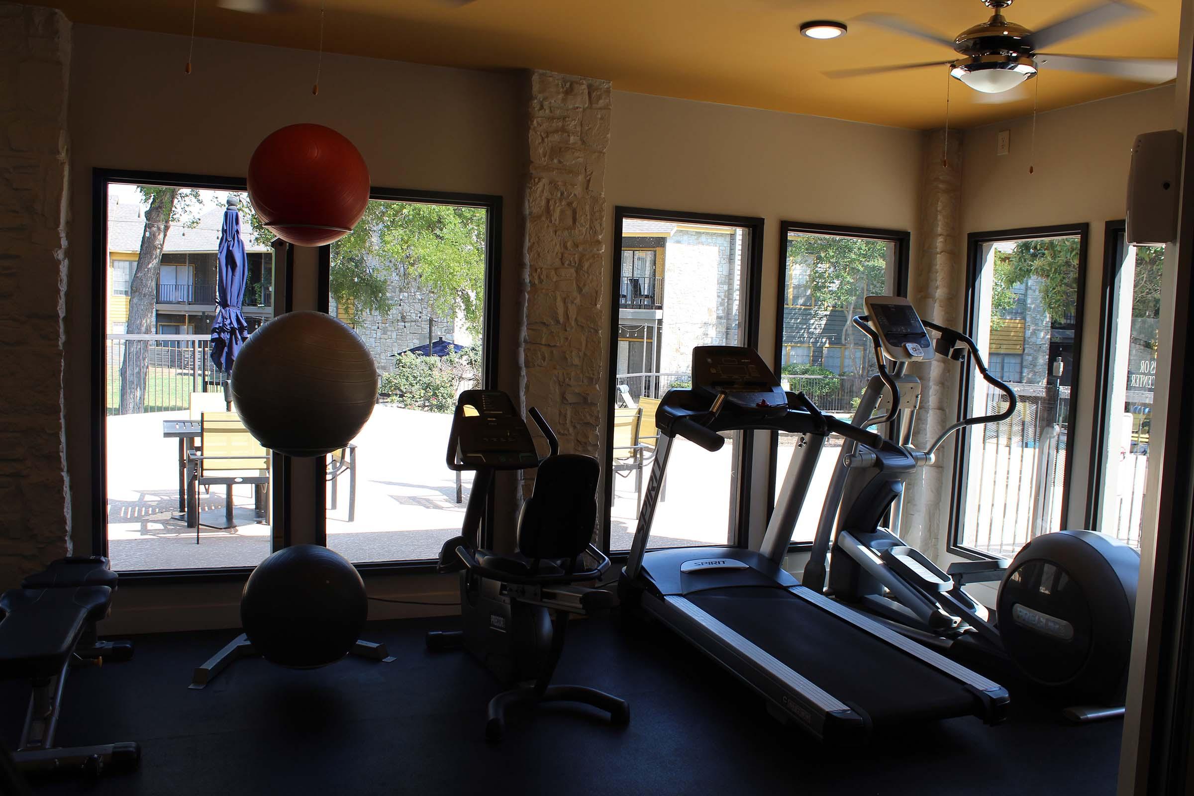 A modern gym interior featuring exercise equipment including treadmills, stationary bikes, and weights. Large windows provide natural light, and exercise balls are arranged decoratively. The view outside shows a patio area with outdoor seating and trees.