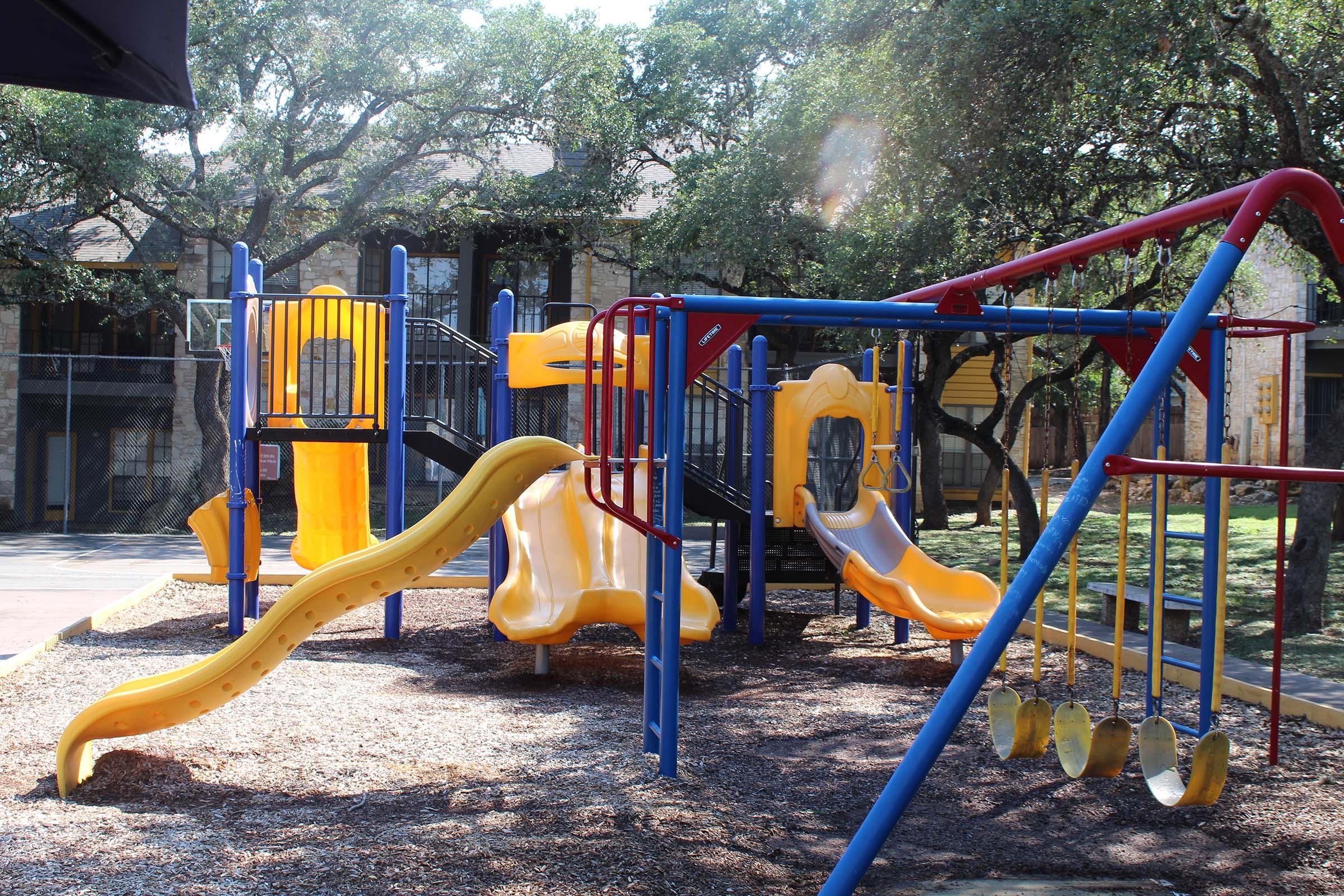 A brightly colored playground featuring yellow slides, climbing structures, and swings, set in a shaded area with trees in the background. The playground is surrounded by wood-chip safety surfacing, providing a fun space for children to play.